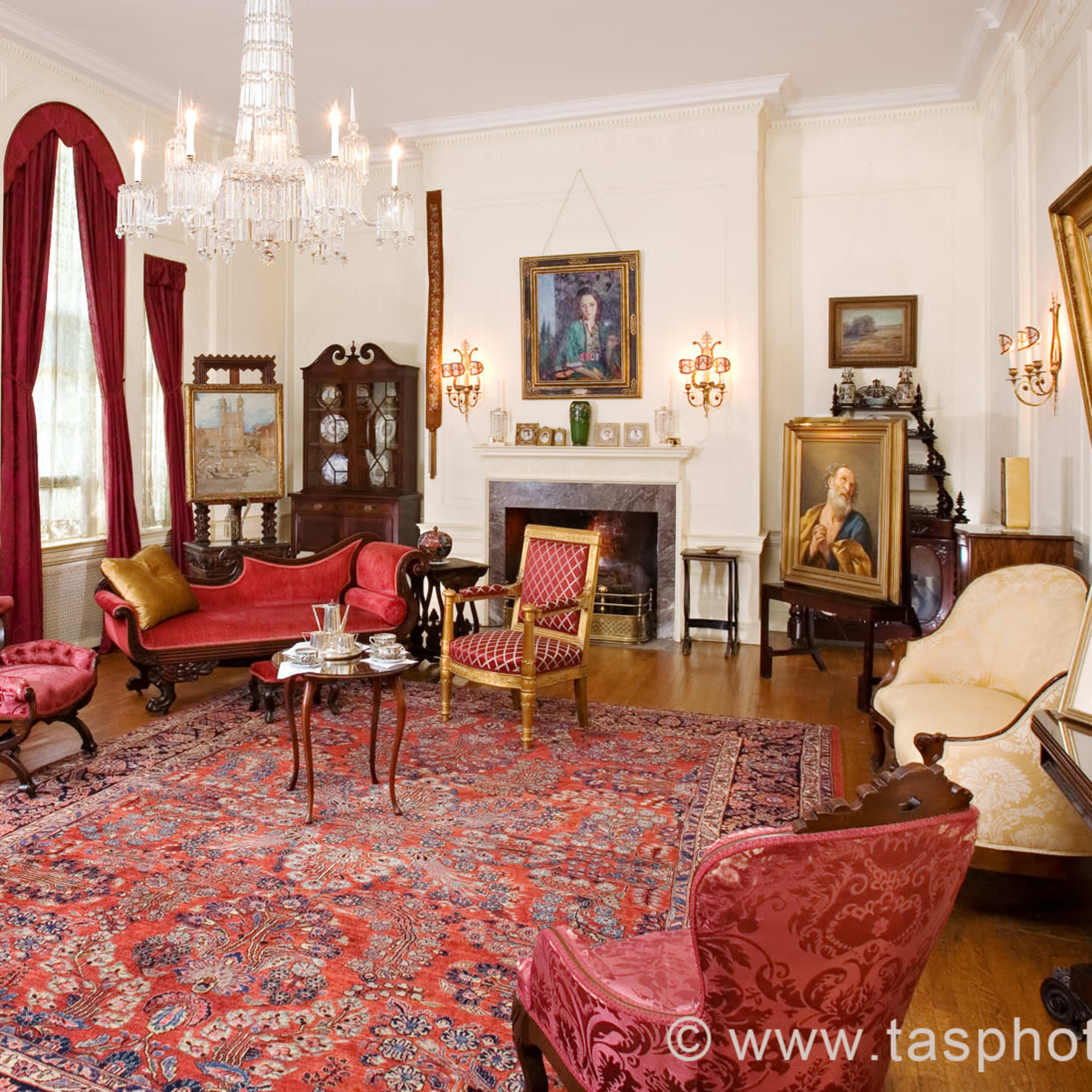 The scene shows a vintage living room with ornate furnishings, including a grand piano, multiple paintings on the walls, and a large, patterned rug.
