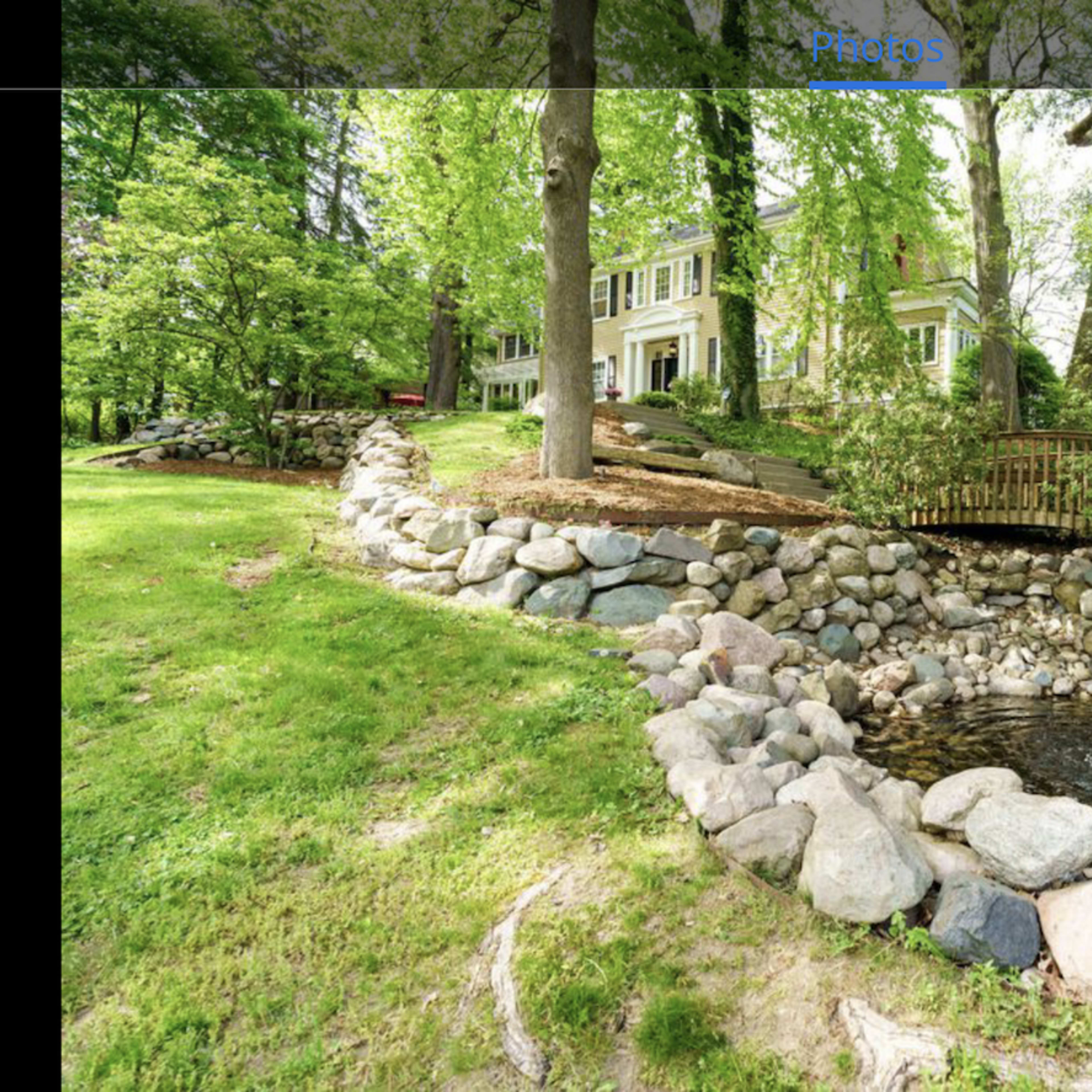 A landscaped yard with a stone-bordered pond and fountain, surrounded by trees and a house in the background.