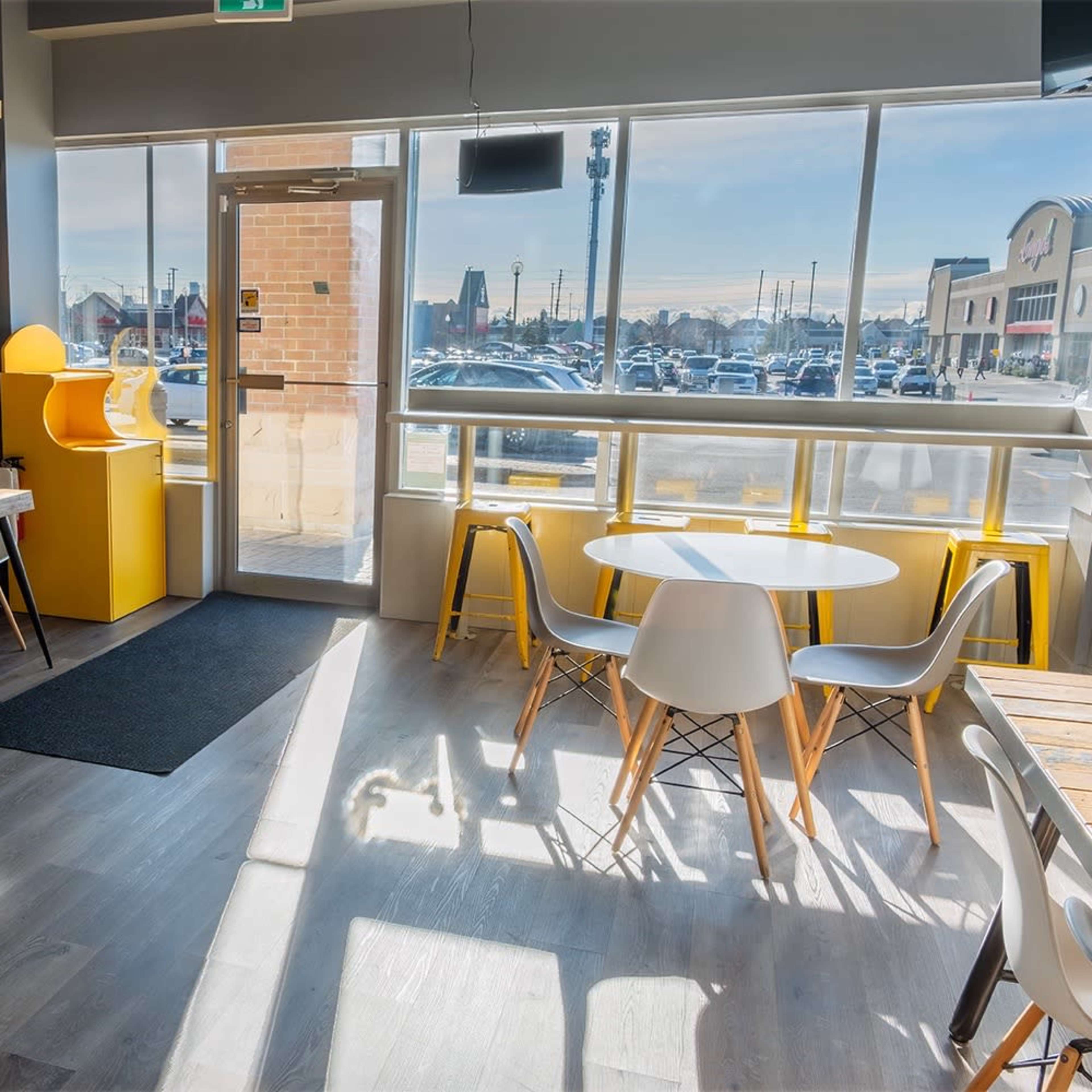 A modern restaurant interior with wooden tables, white chairs, and large windows that provide natural light, featuring a large pizza mural on one wall.