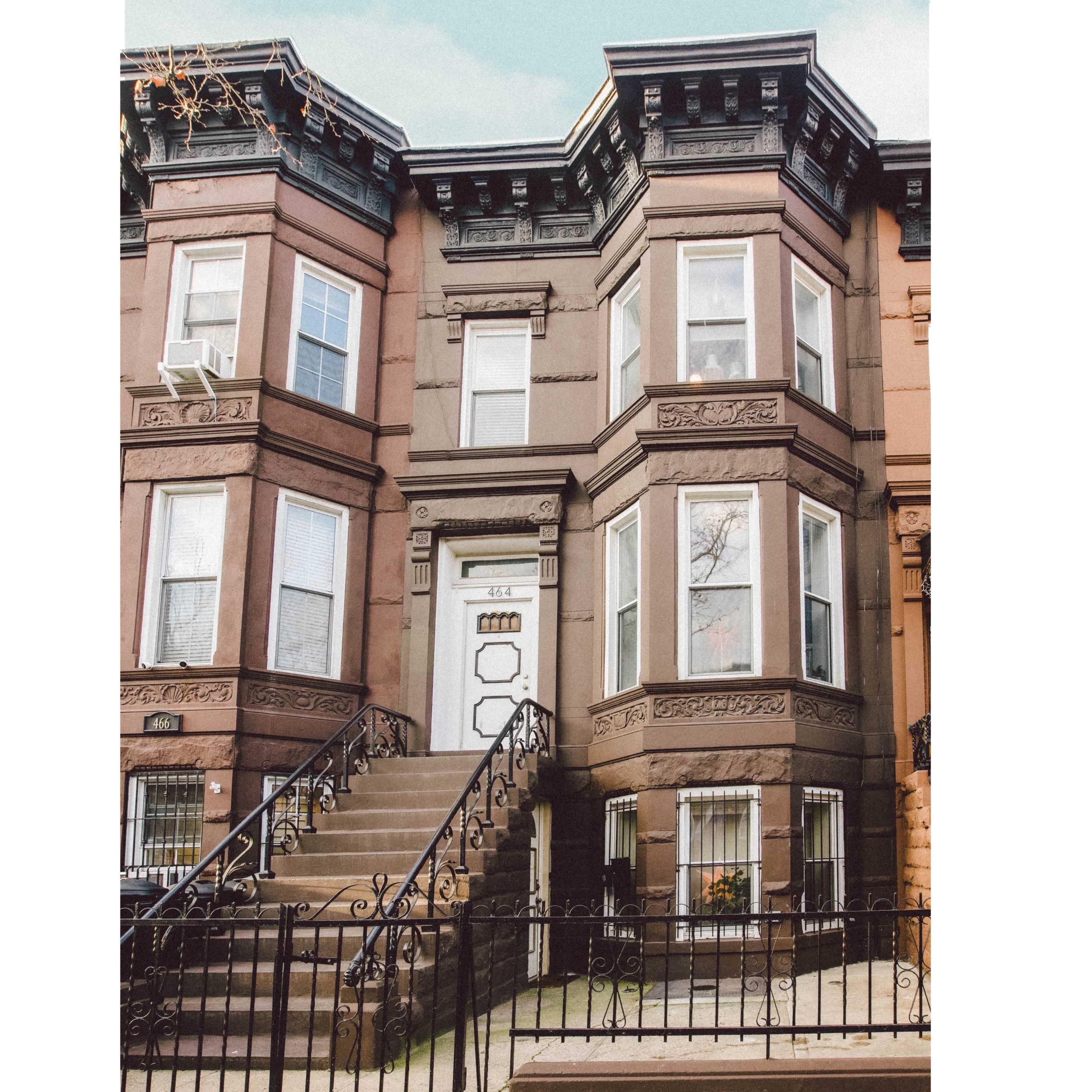 A brownstone building with a set of stairs leading to a door framed by large windows on either side.