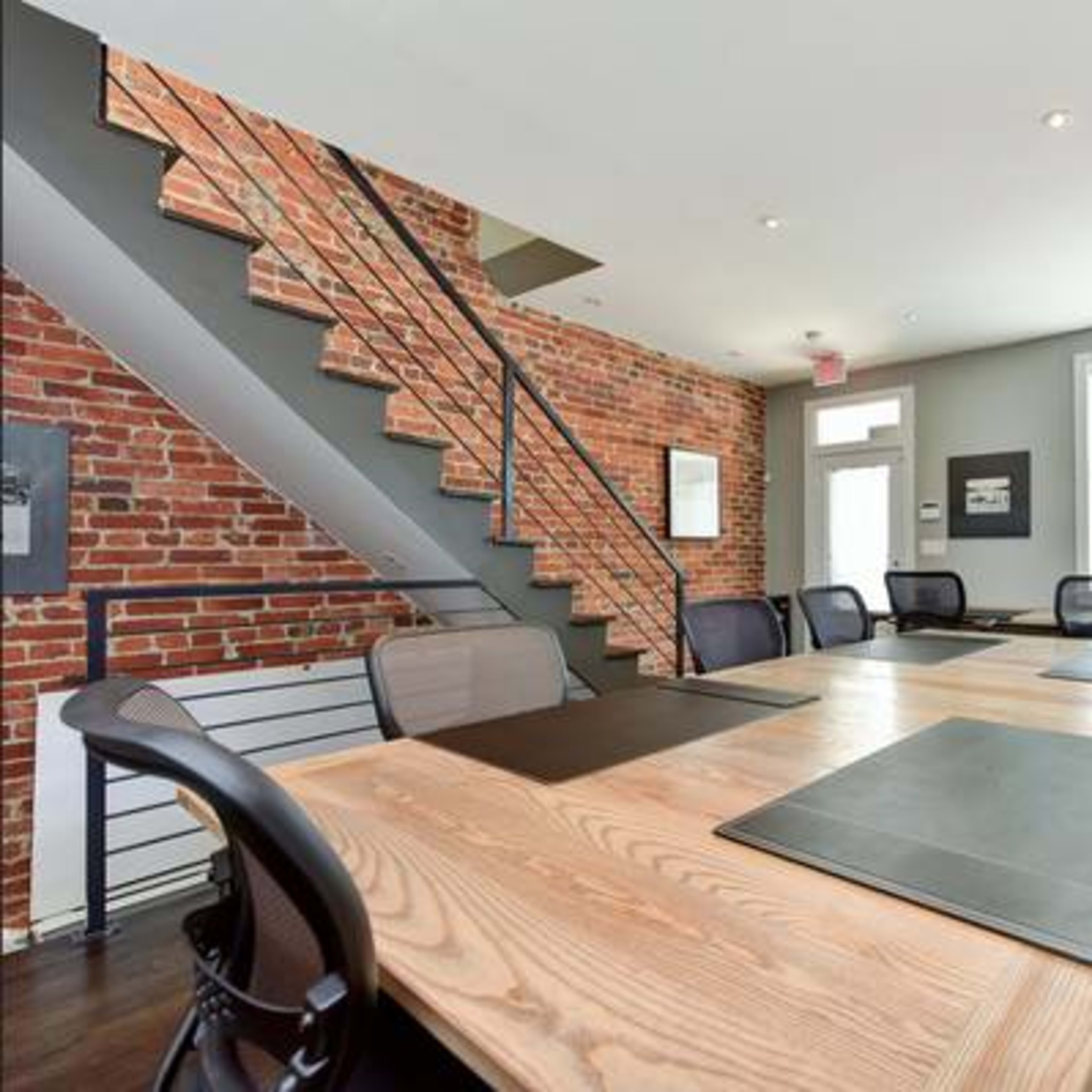 A modern meeting room with a large wooden table surrounded by black chairs, a red brick wall, and a staircase in the background.
