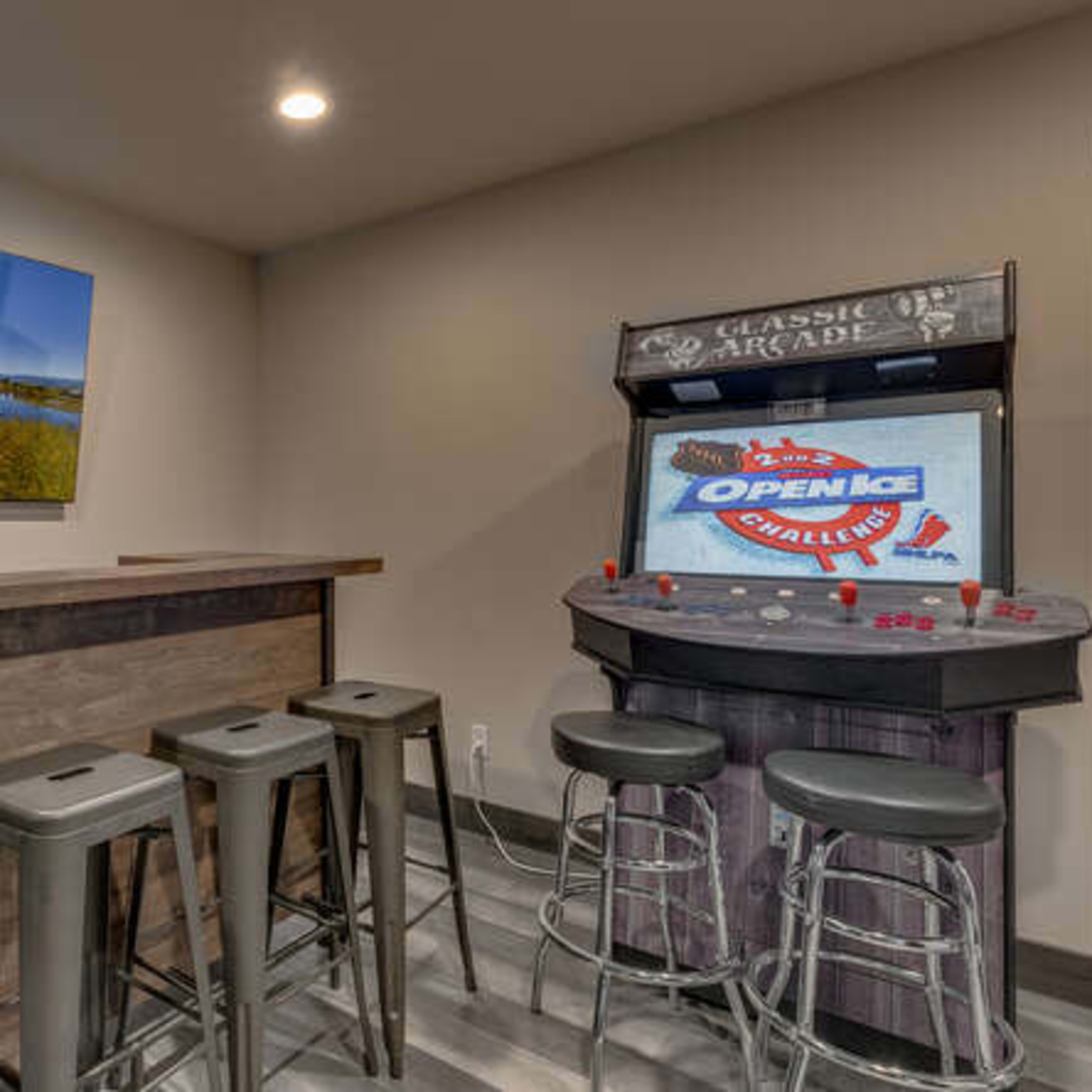 A room with a dark wooden bar and four metal stools, featuring an arcade game machine against the wall with a large television displaying a landscape photograph.