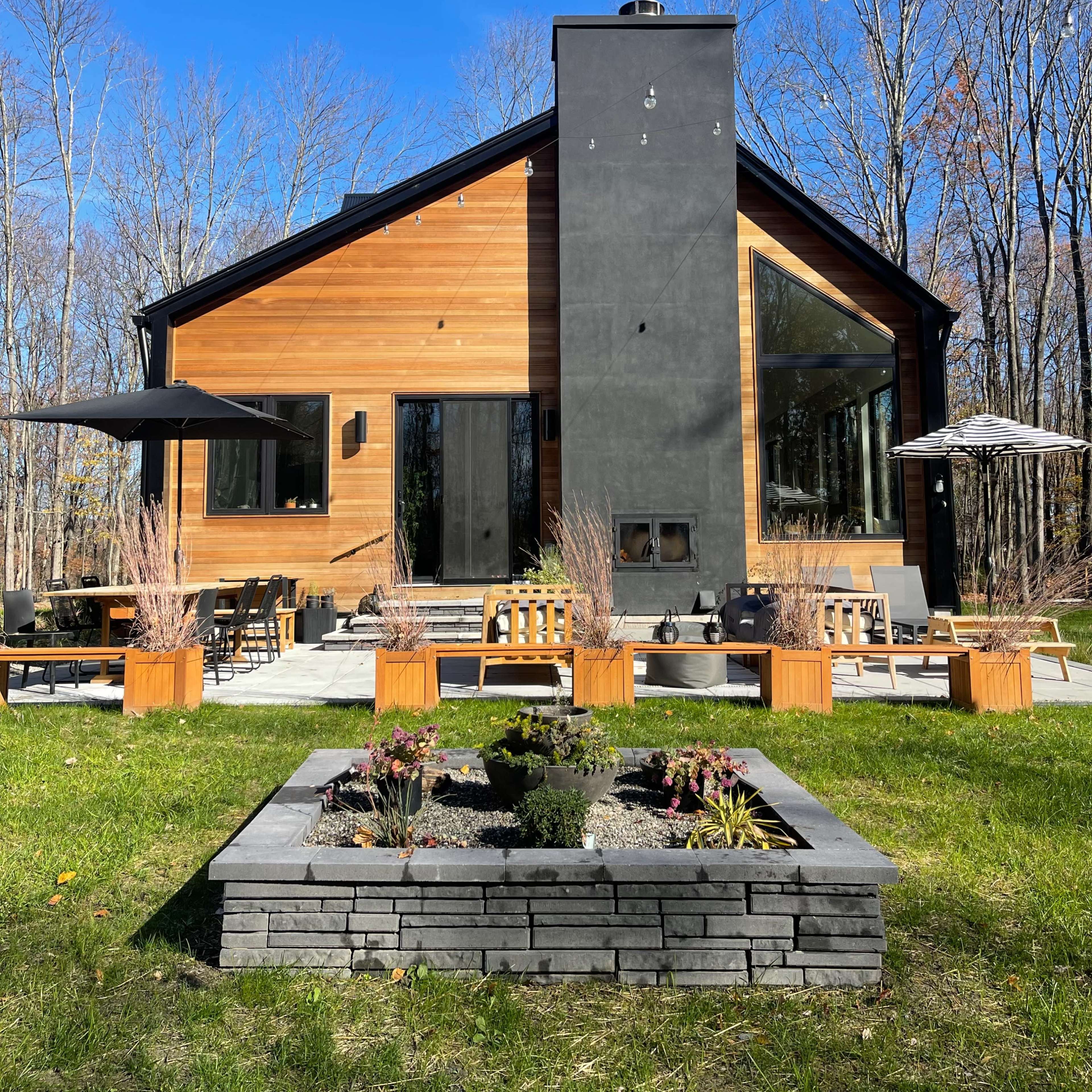 A modern house with a wooden exterior and a prominent black chimney, set against a backdrop of trees and a landscaped garden featuring a stone planter.