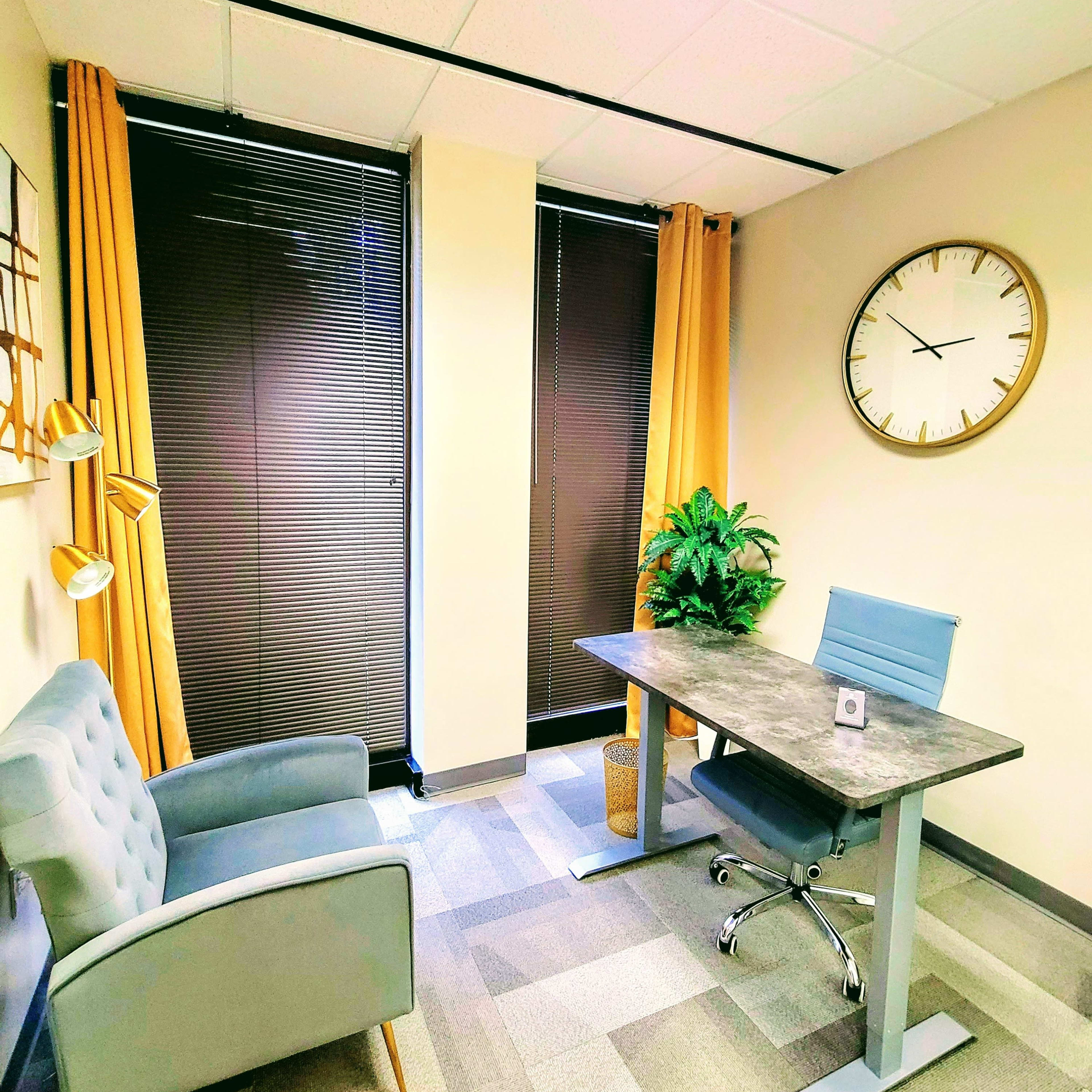 A modern office space featuring a gray desk, a blue chair, a small plant, and a comfortable armchair, with large windows covered by blinds and yellow curtains.