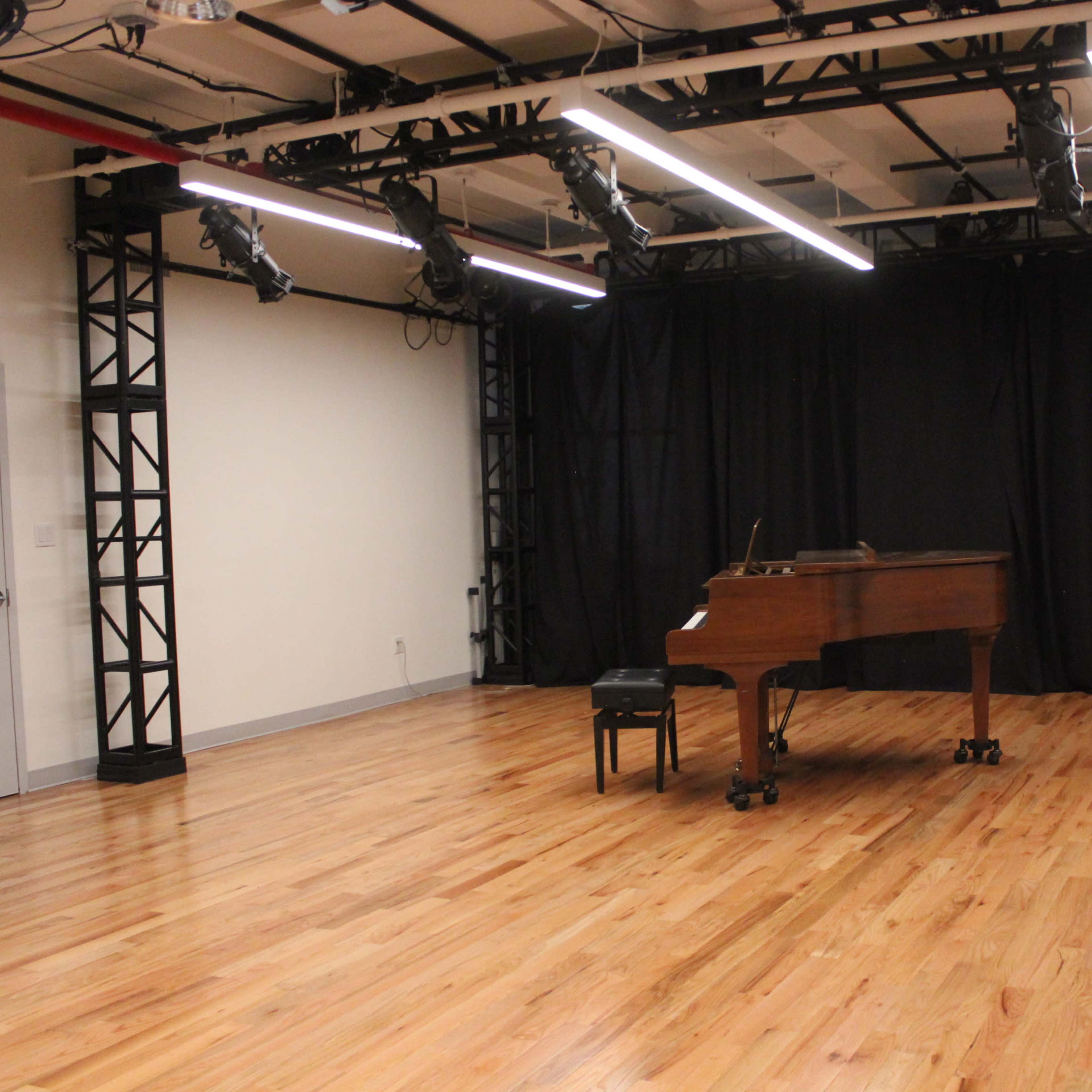 A grand piano and a stool are positioned on a wooden floor in an empty room with black curtains and overhead stage lighting.