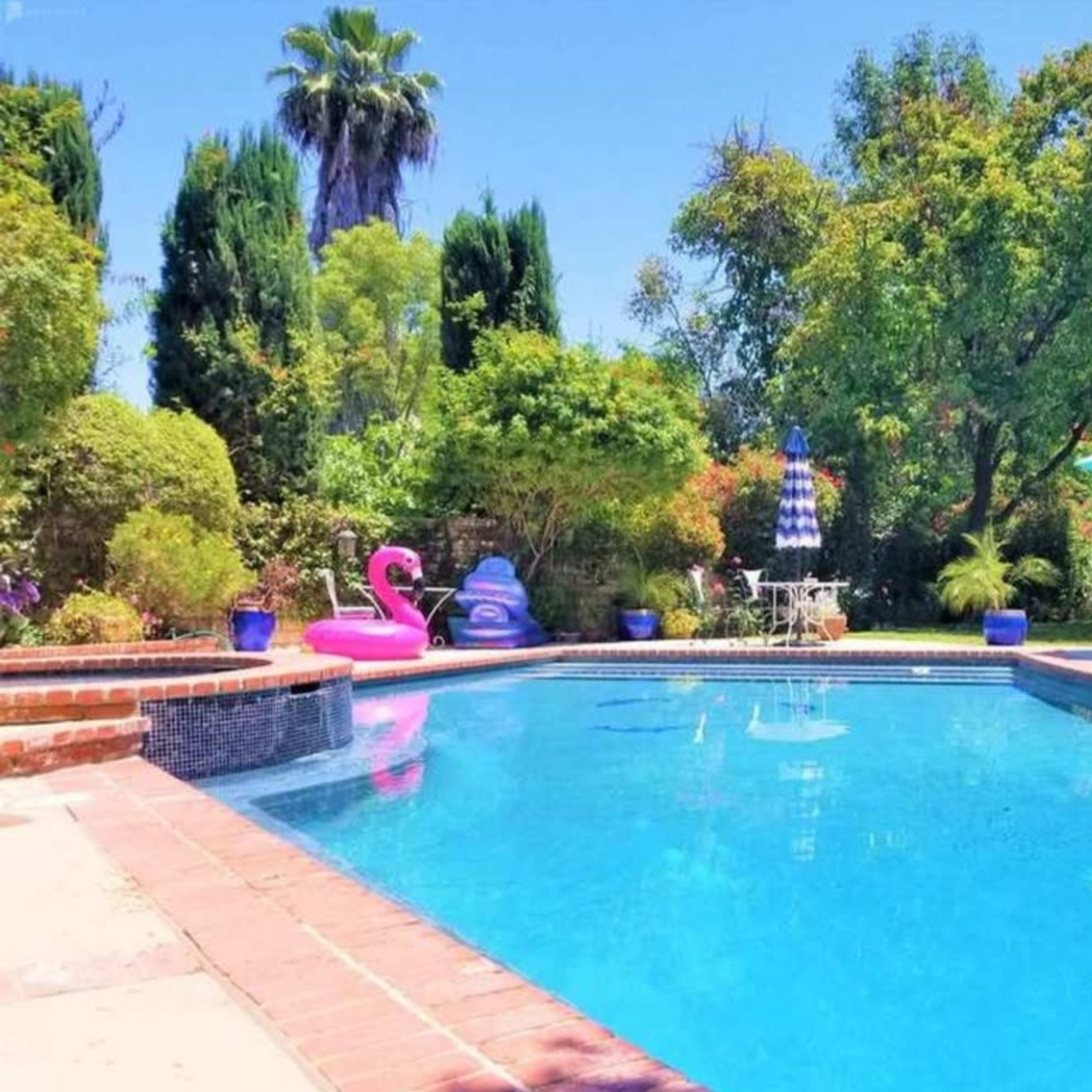 A clear swimming pool is surrounded by green trees, patio furniture, and a pink flamingo float.