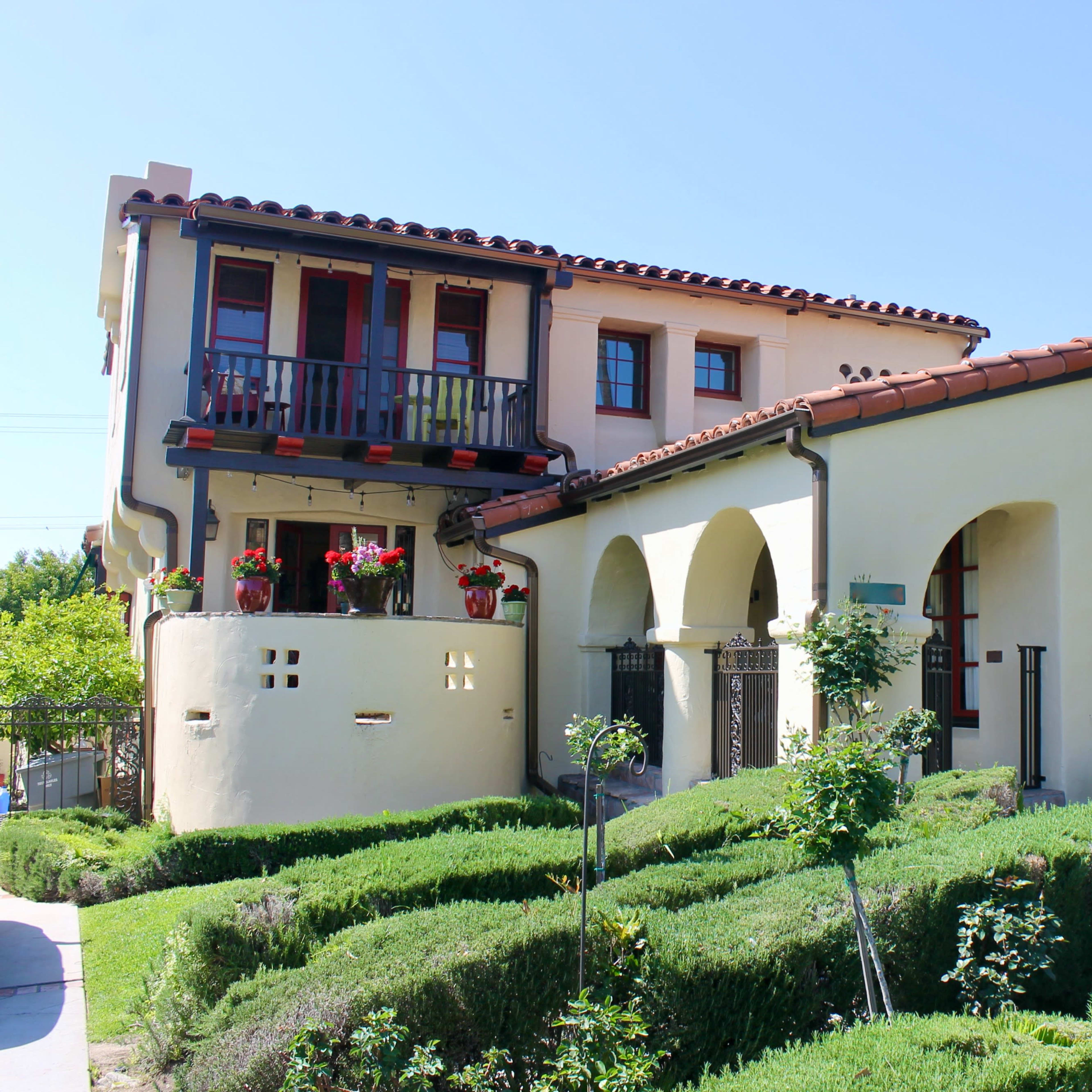 A two-story house with a red-tiled roof, arched windows, and manicured shrubs in the front yard.