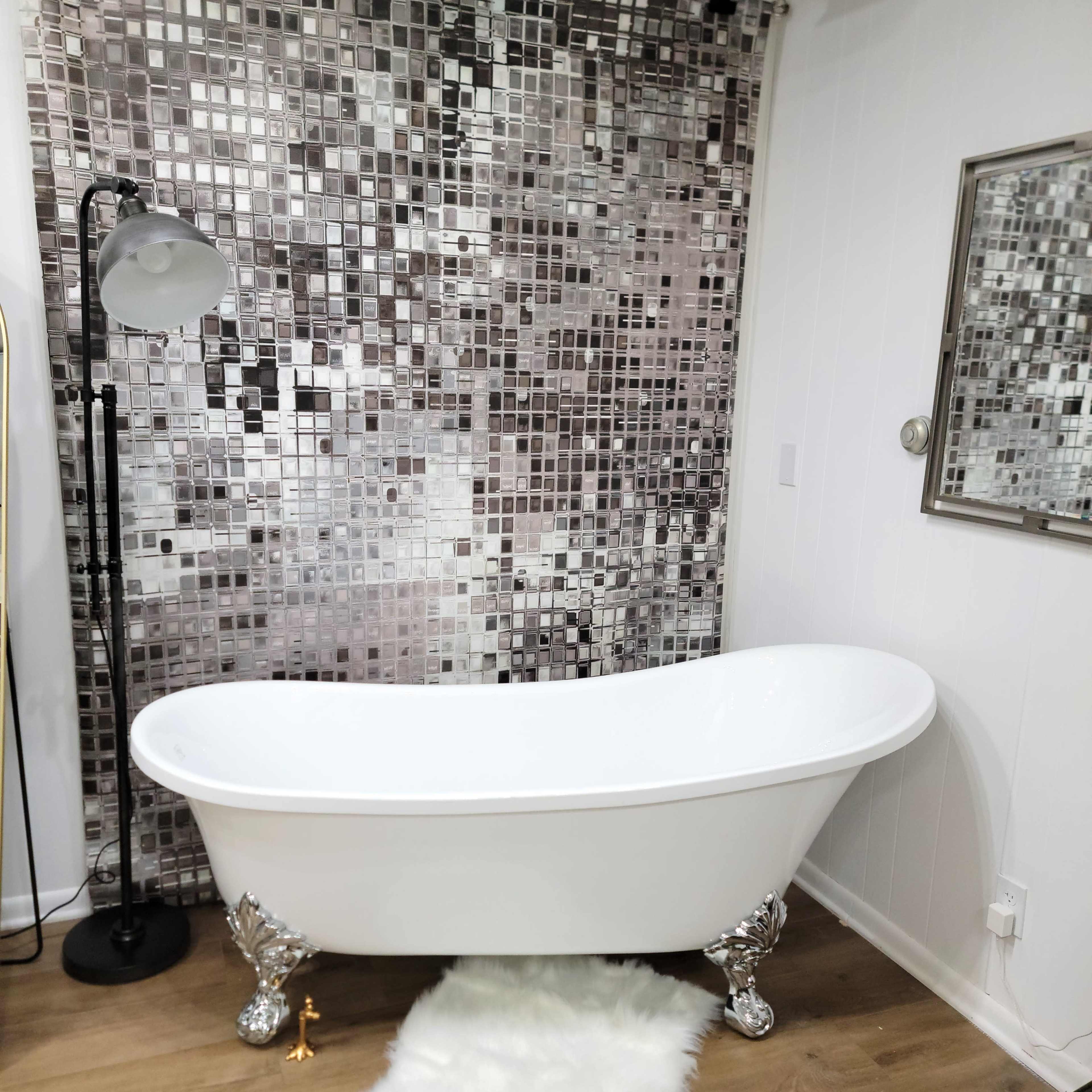 A freestanding white bathtub with ornate silver legs is placed on a light wooden floor in front of a reflective mosaic tile wall and a mounted mirror.