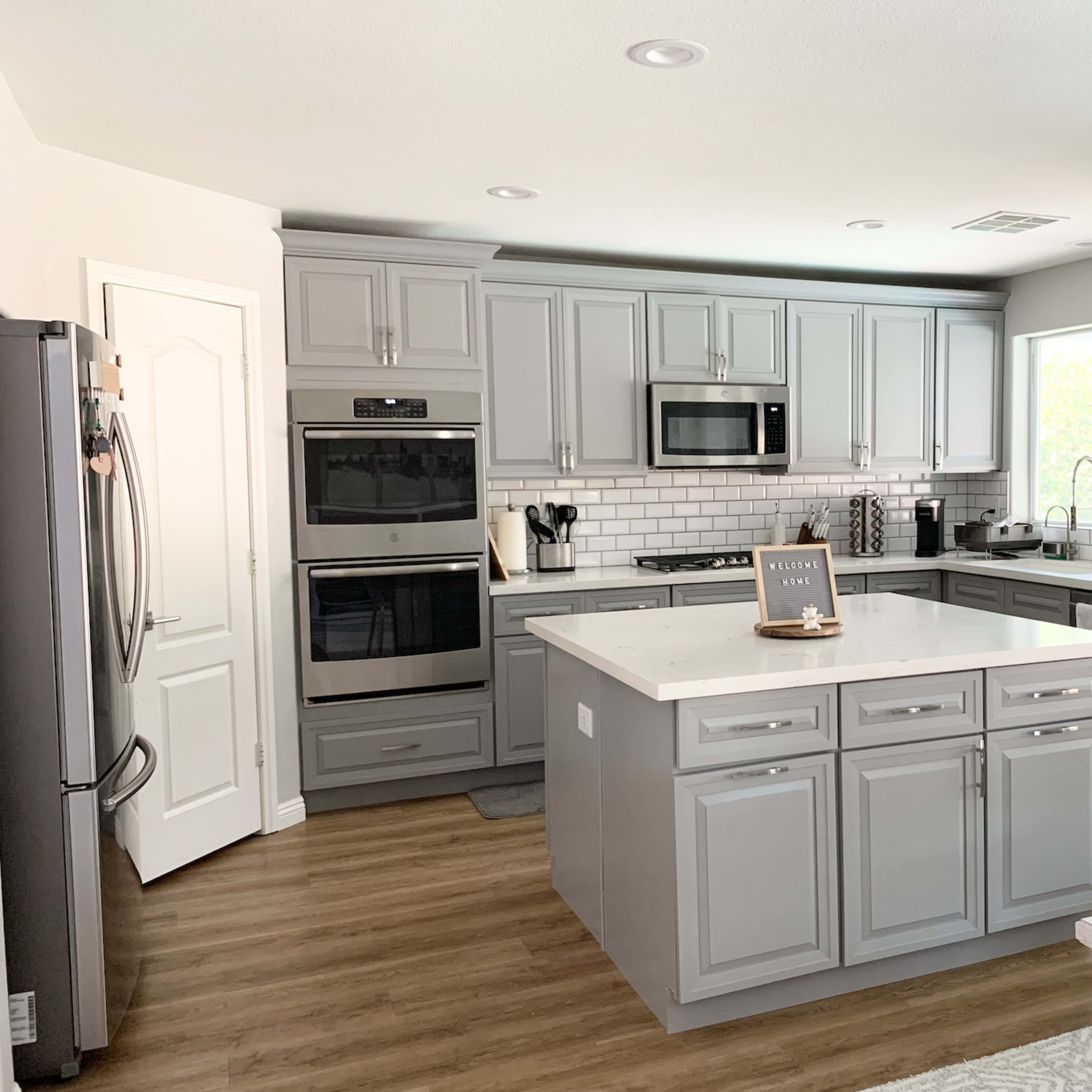 The kitchen features gray cabinets, stainless steel appliances, a central island with seating, and a window providing natural light.
