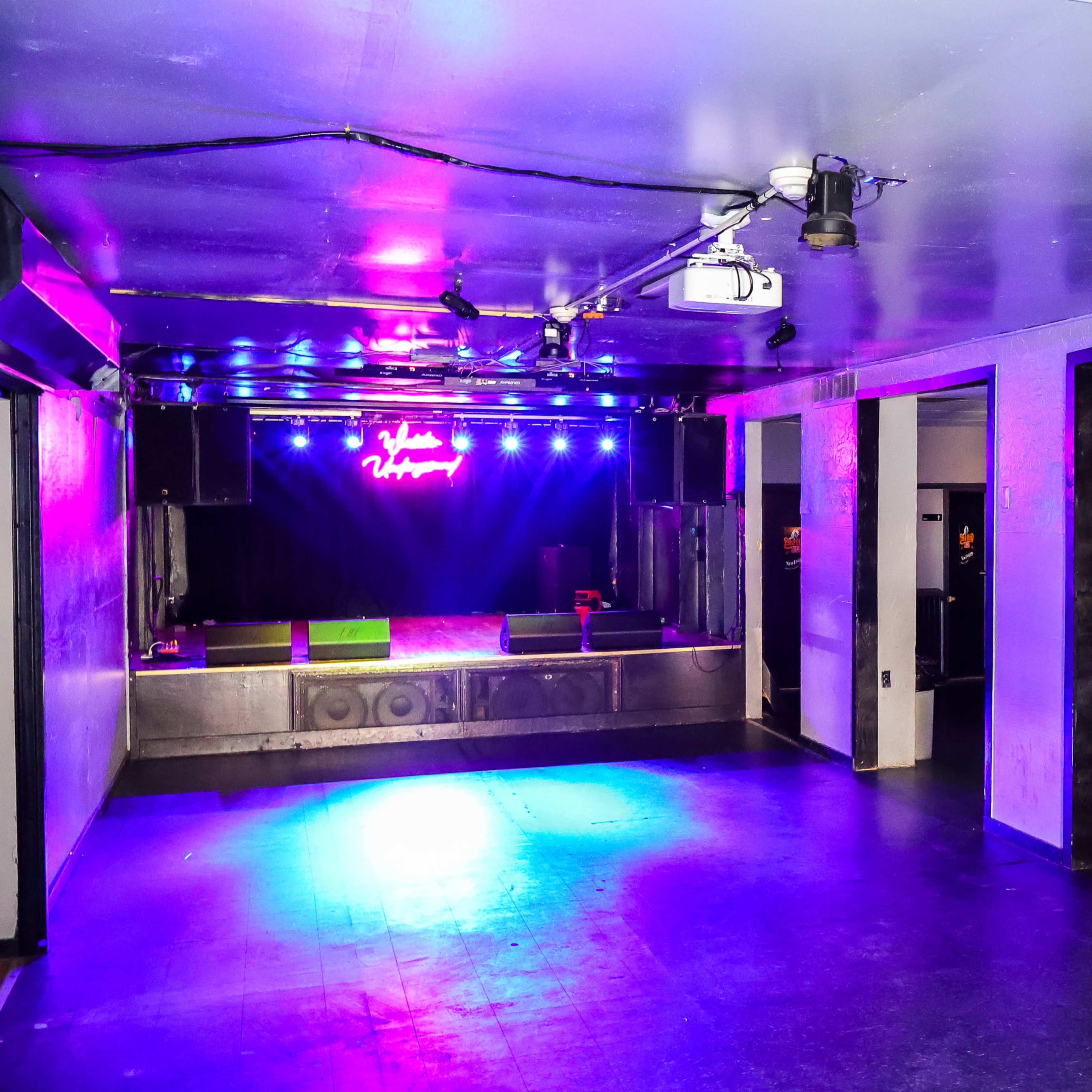 An empty music venue with a stage illuminated by colorful lights and a bar area in the background.