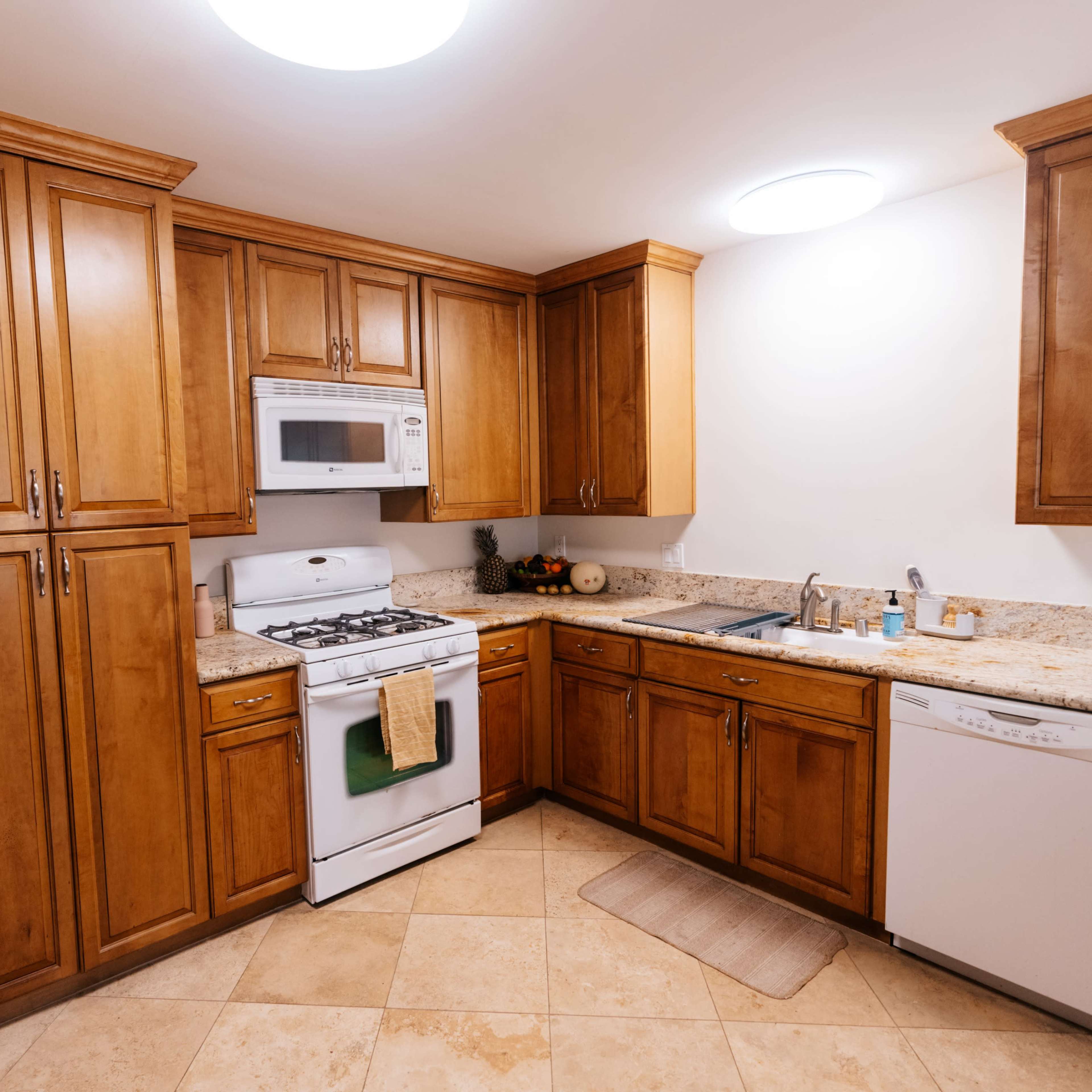 A modern kitchen with wooden cabinetry, a white stove and microwave, and beige tile flooring.