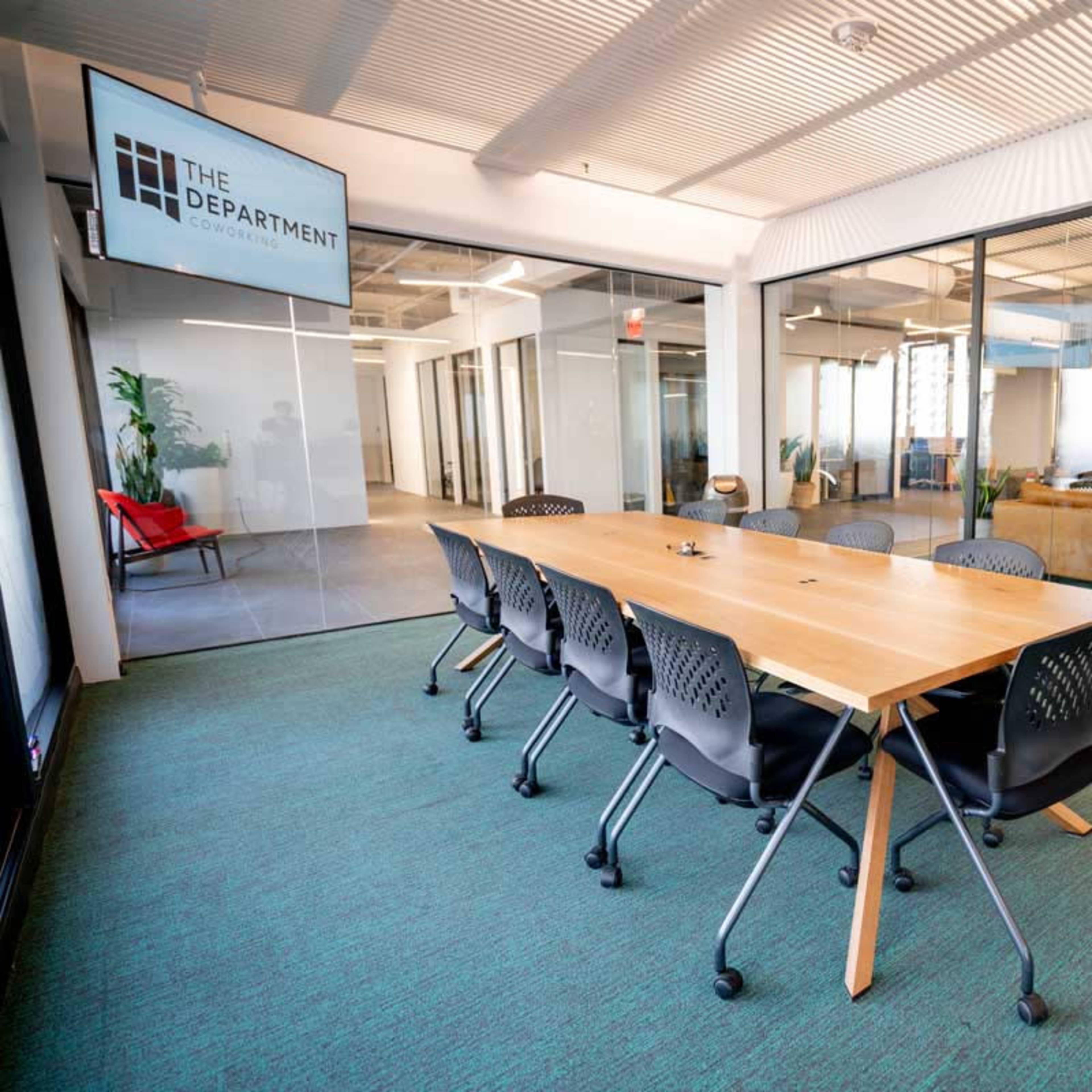 A long wooden conference table with rolling chairs is situated in a modern office space featuring glass walls and green carpeting.