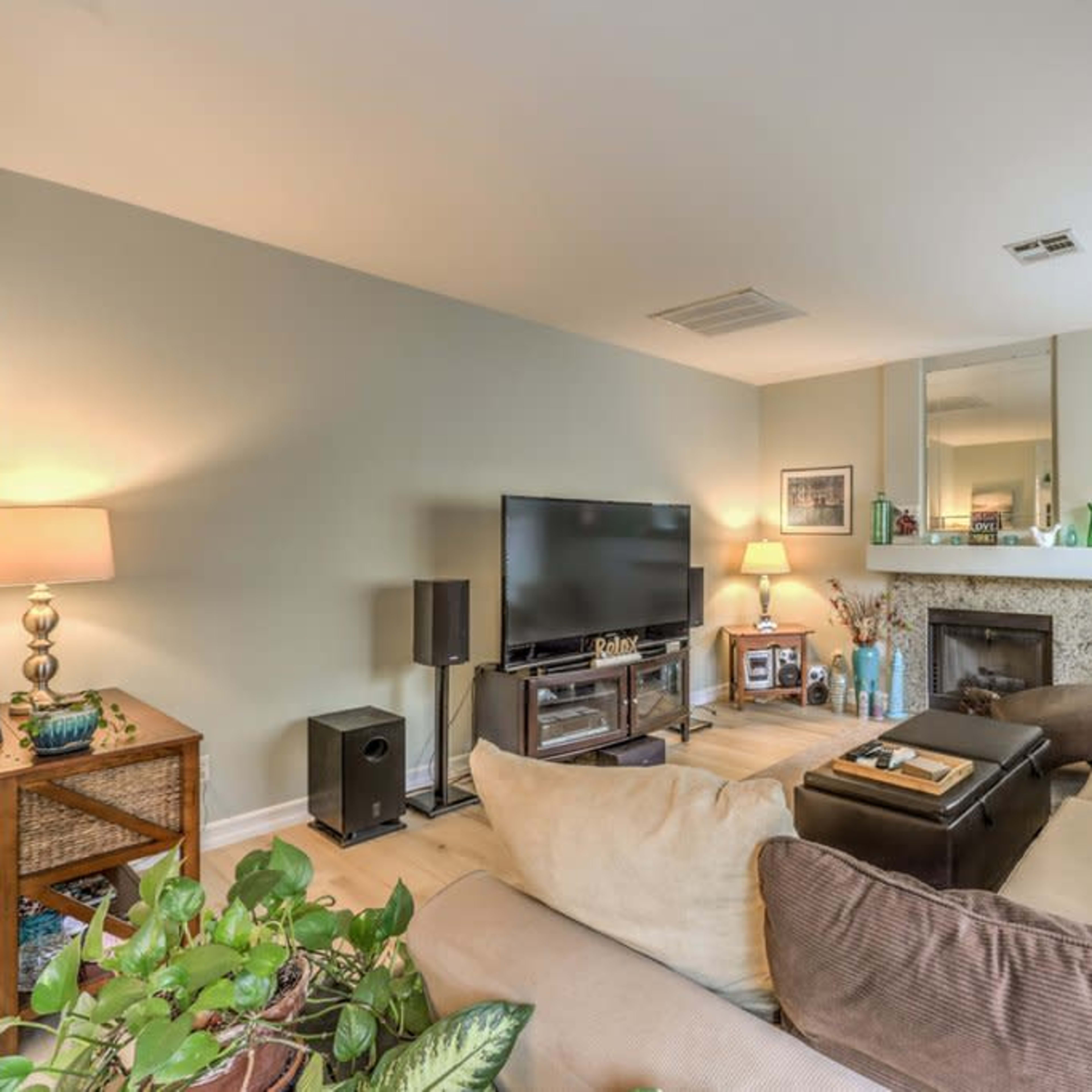 A living room features a sectional sofa, a television stand, a fireplace, and decorative elements including plants and framed pictures.