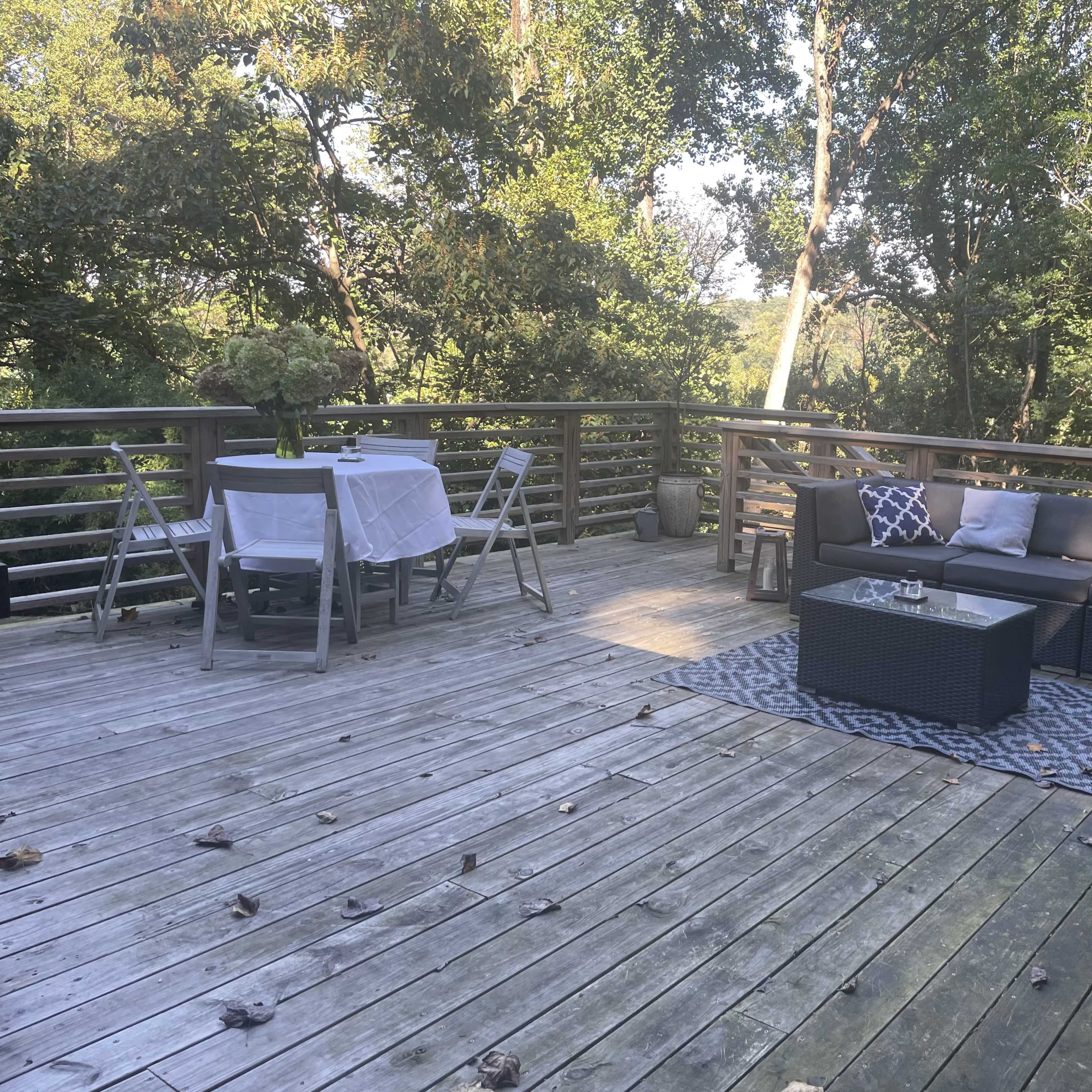 A wooden deck furnished with a sectional sofa, a coffee table, and dining setup, surrounded by trees.