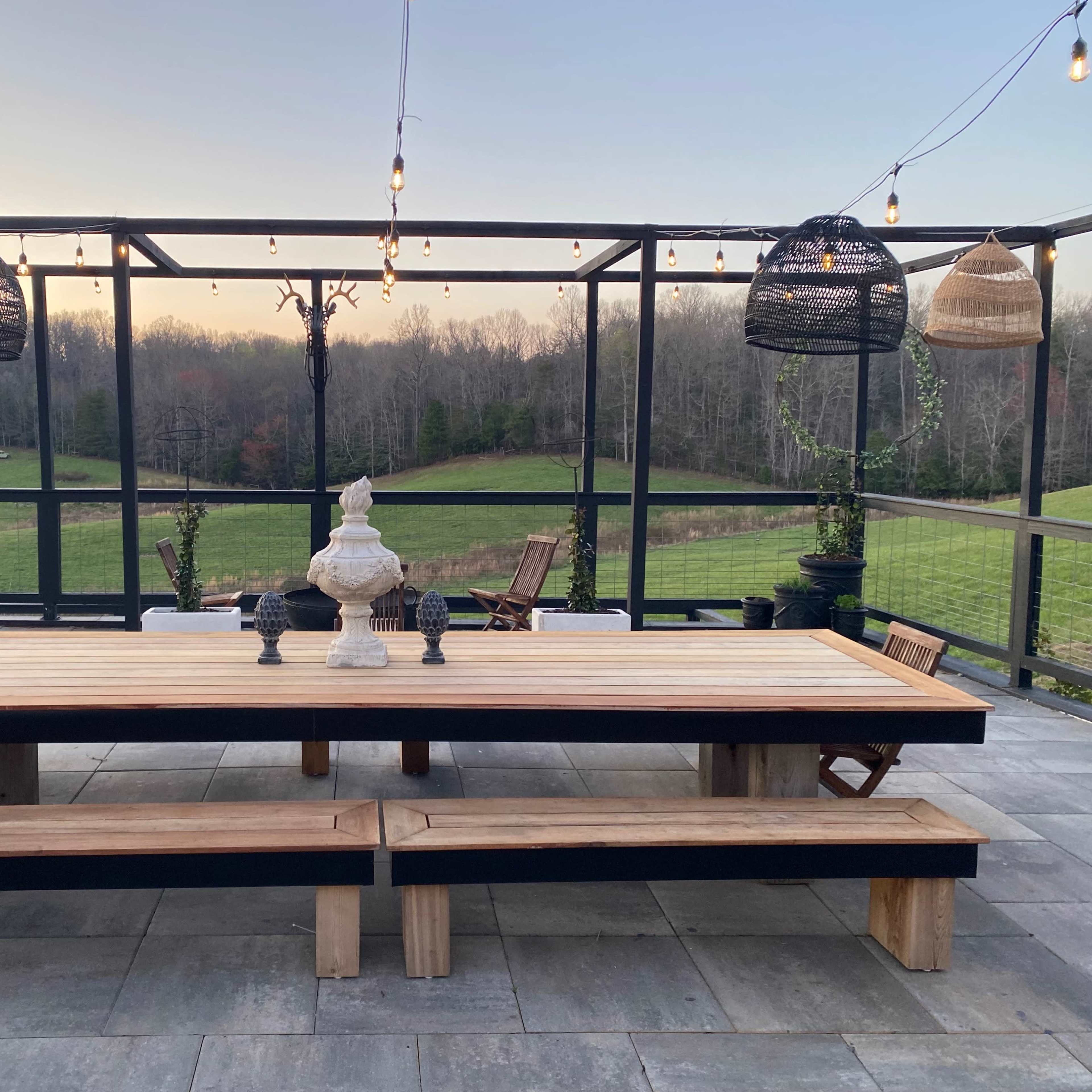 A spacious outdoor dining area features a large wooden table, benches, and decorative pendant lights, set against a backdrop of a green field and forested area.