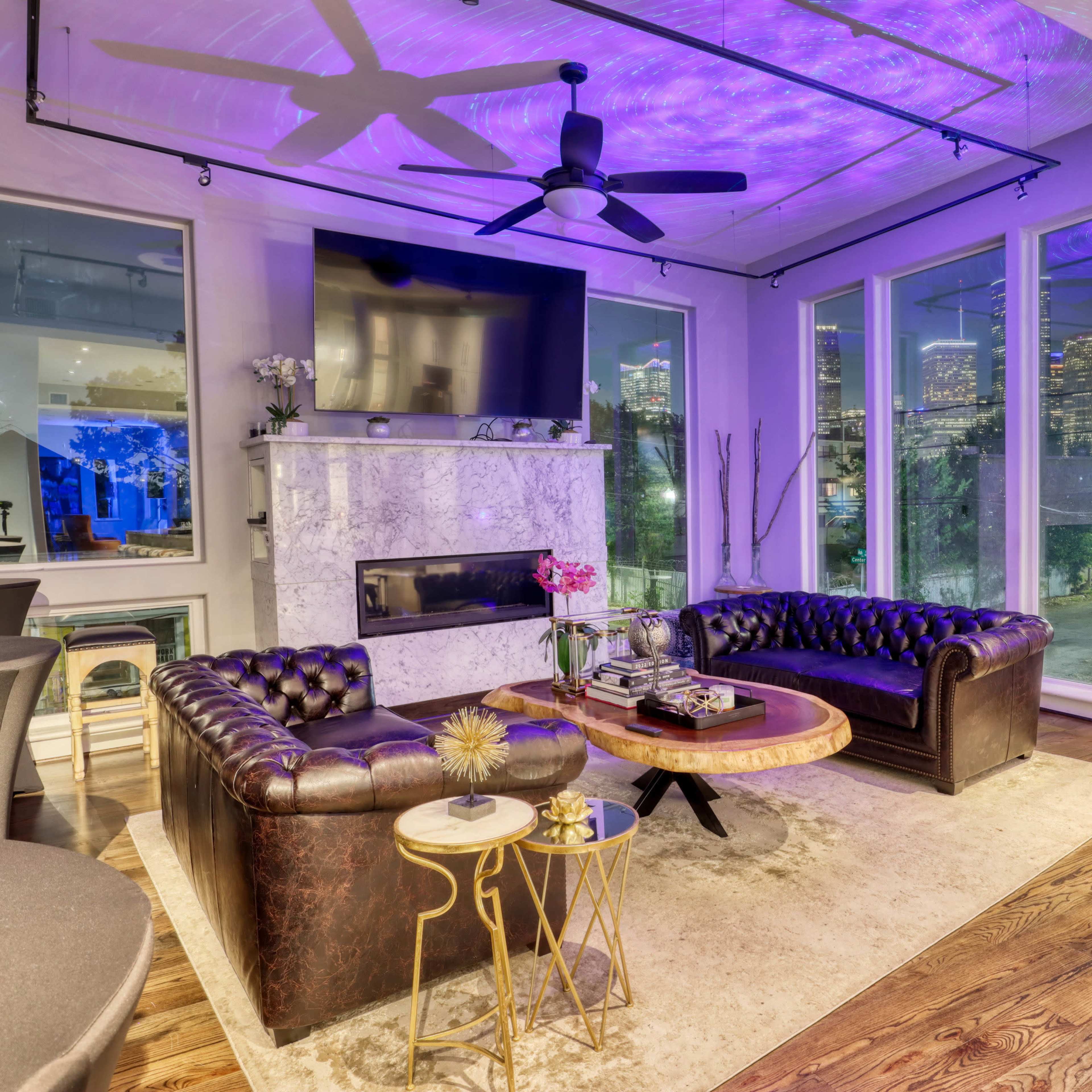 A modern living room with a marble fireplace, two leather sofas, a round coffee table, and large windows offering a view of a city skyline.