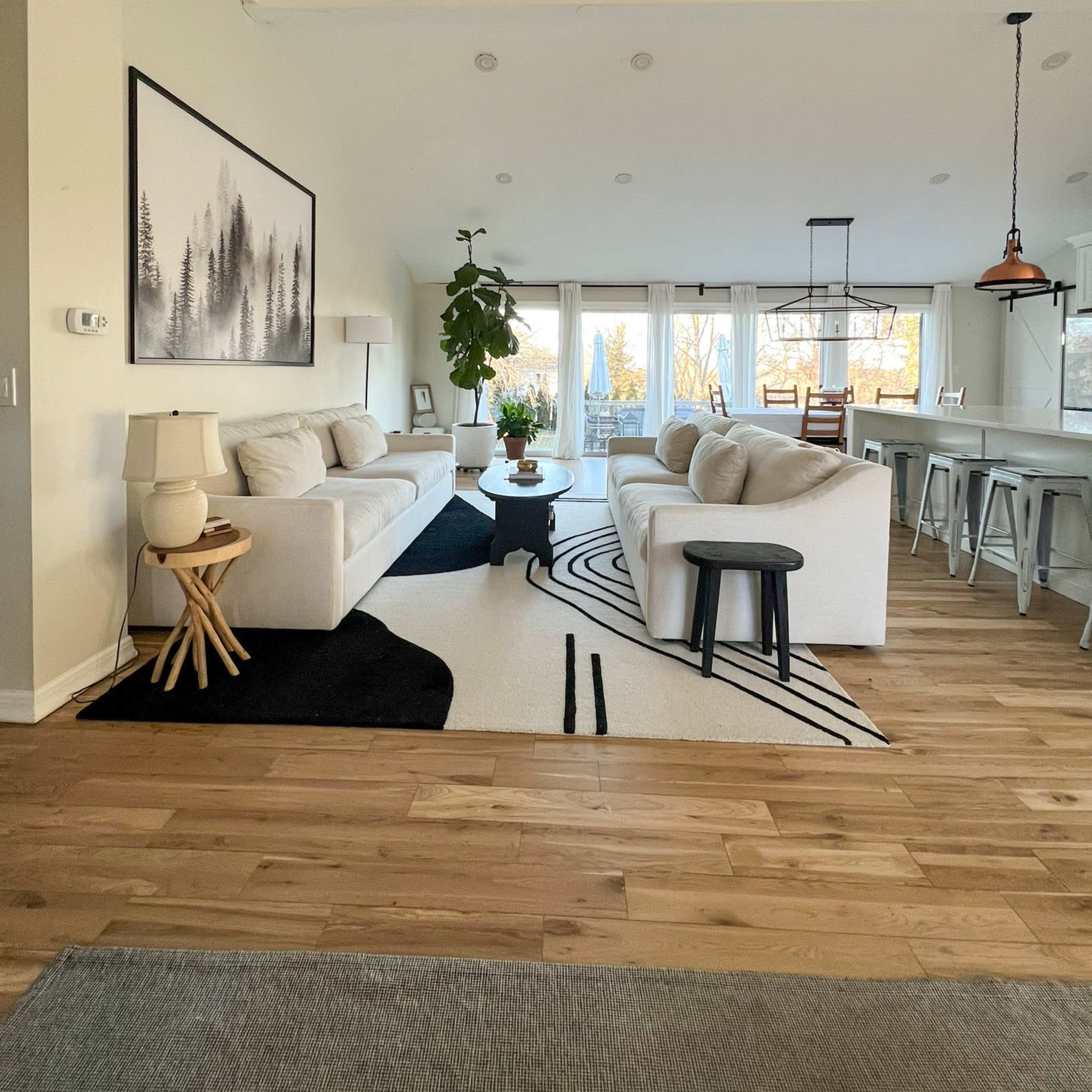 A spacious living area features two white sofas facing each other, paired with a round black coffee table, a large rug with a geometric design, and a dining area visible through large windows.