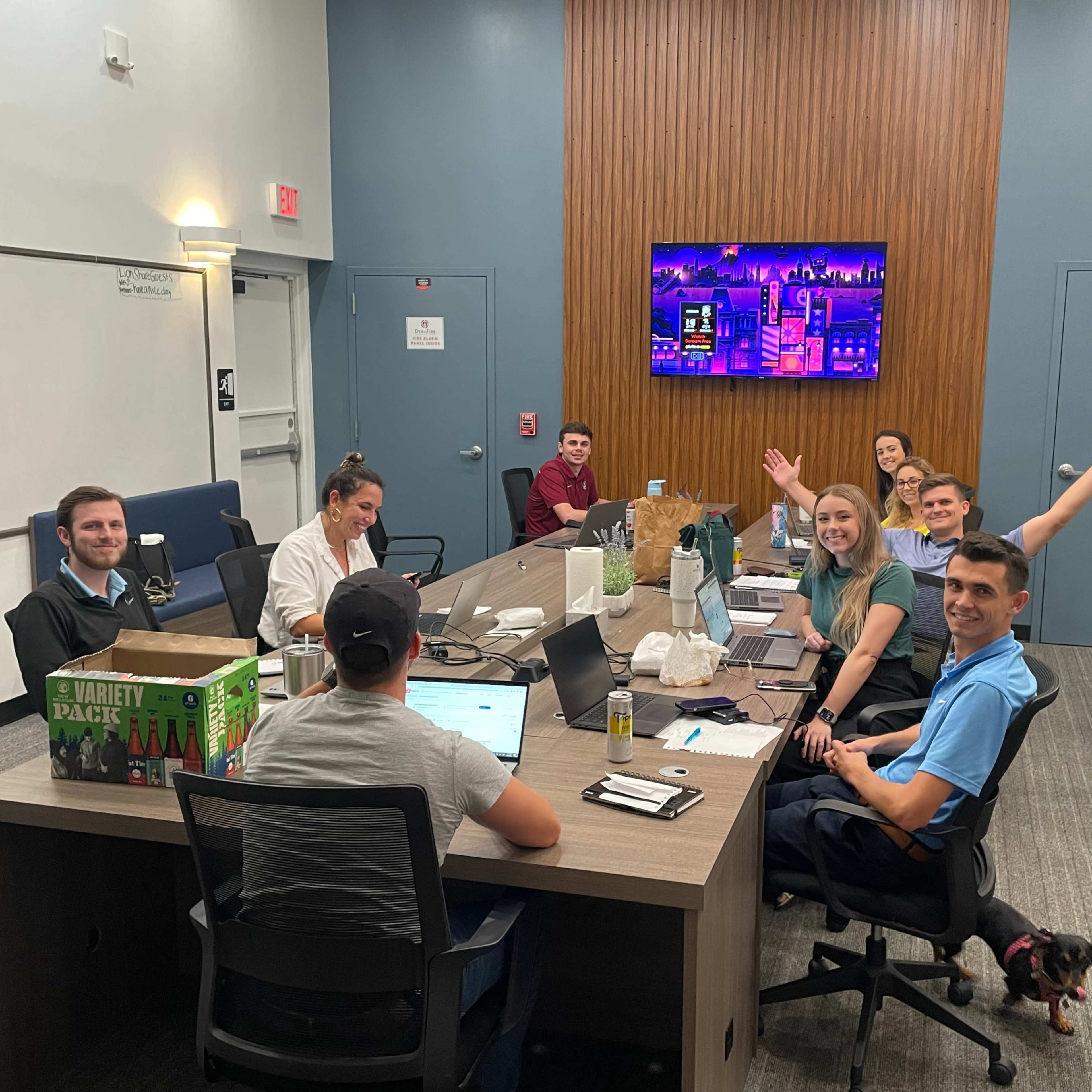 A group of eight people is seated around a conference table in a meeting room with laptops and a large screen displaying colorful content in the background.