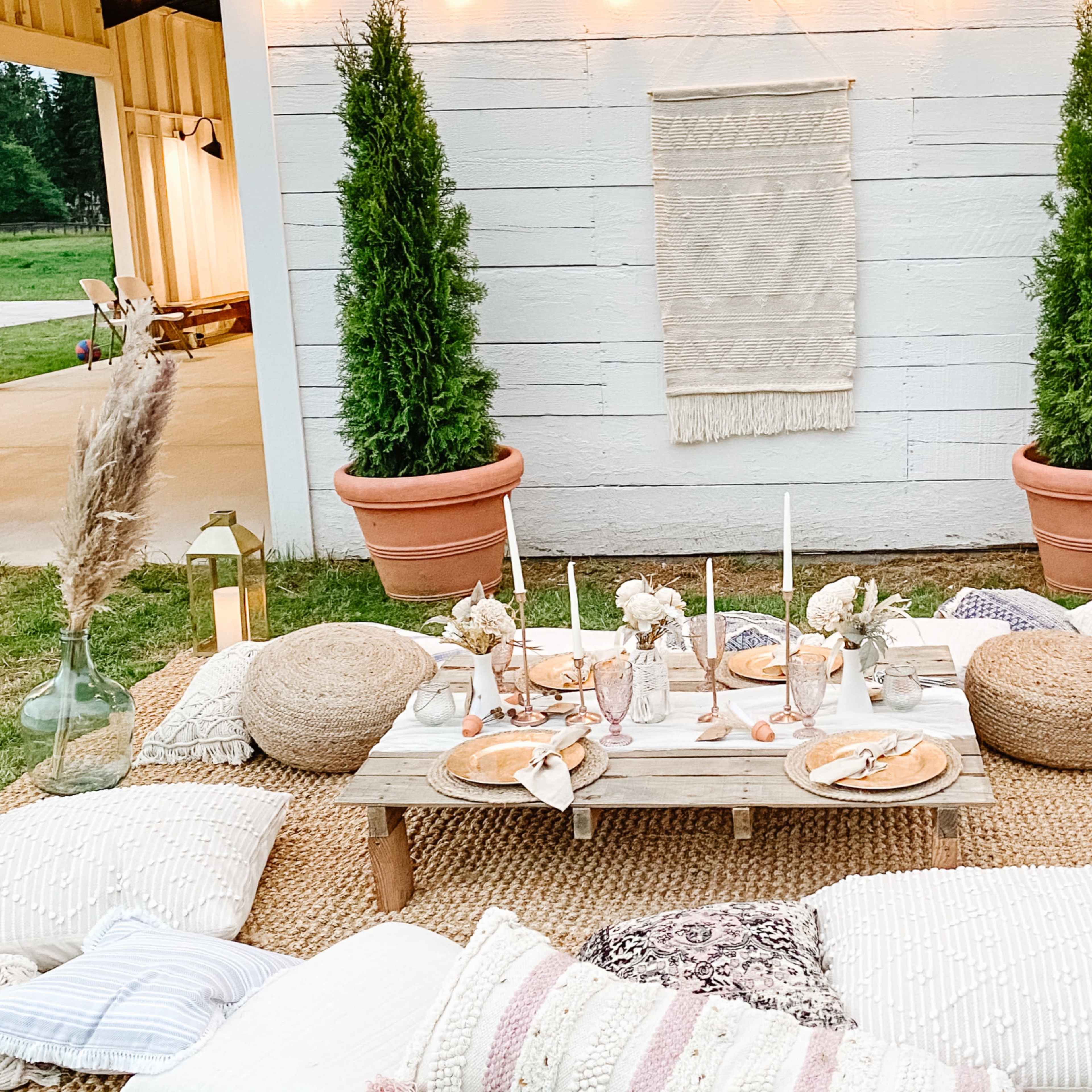 An outdoor gathering area with a low wooden table surrounded by cushions and decorative items, set against a backdrop of potted plants and soft lighting.