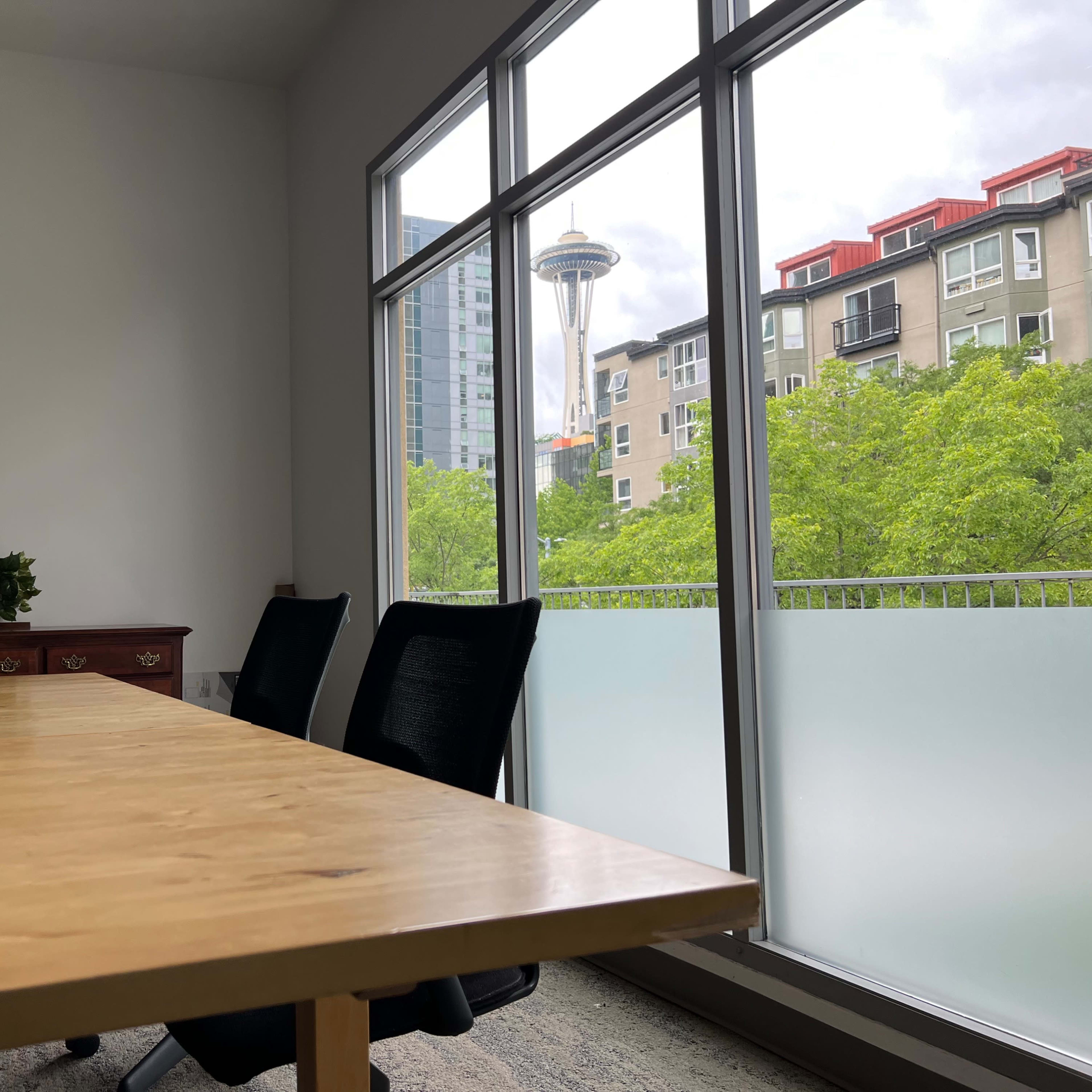 A wooden meeting table with black chairs is positioned in a conference room, overlooking green trees and the Seattle Space Needle through large windows.