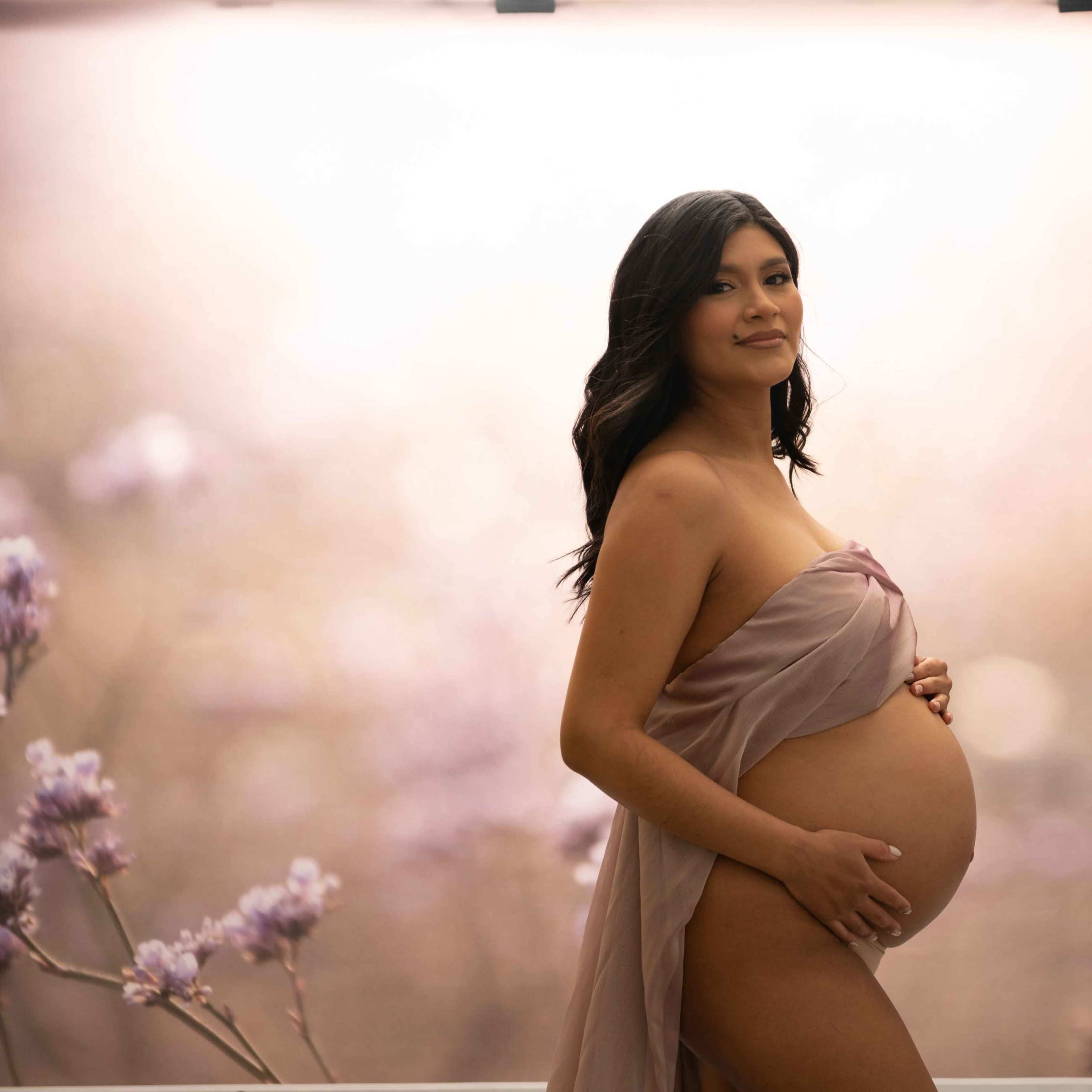 A pregnant woman poses gracefully, holding her belly, against a soft, floral backdrop.