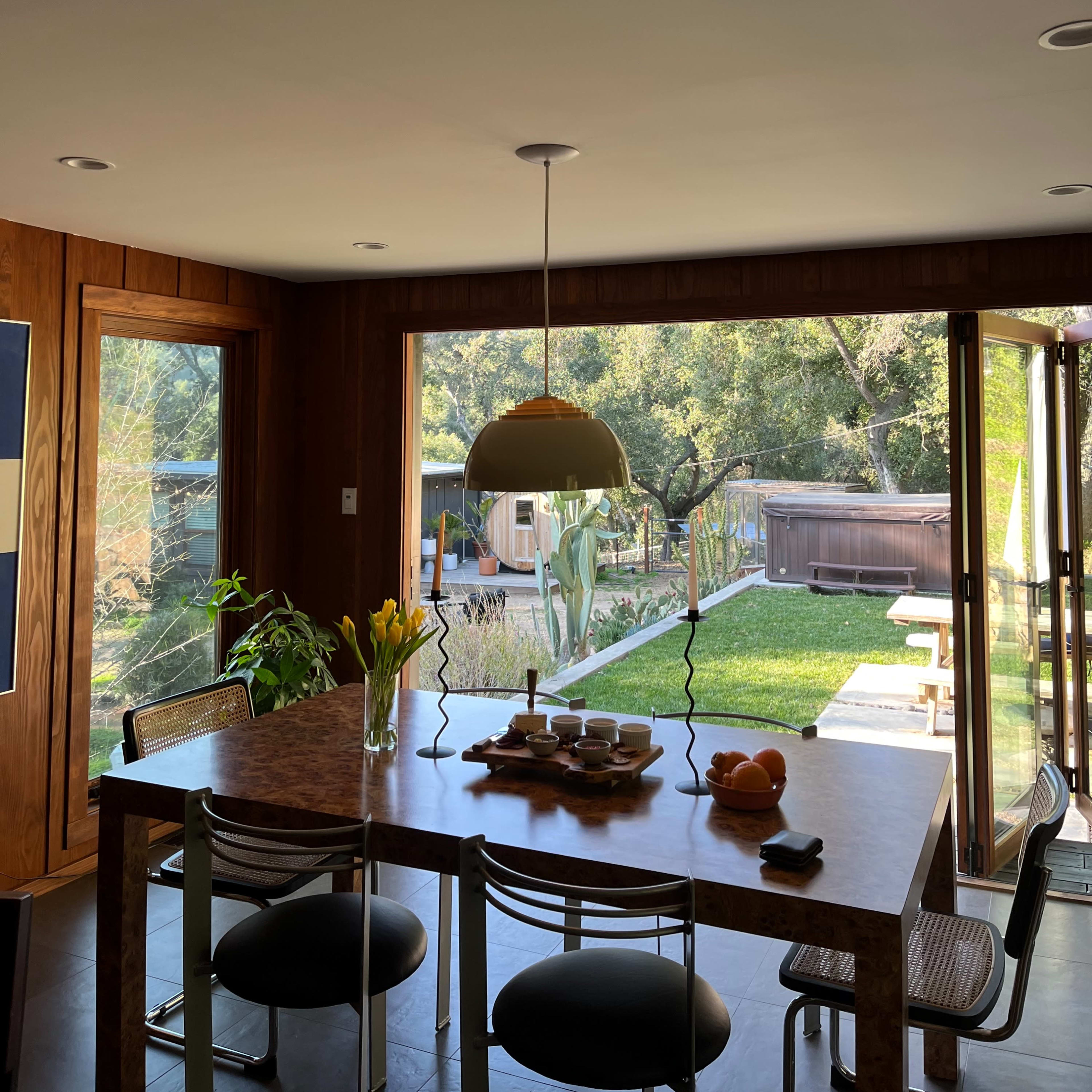 A dining area features a wooden table with a pendant light overhead, surrounded by chairs, and large sliding glass doors open to a green backyard.