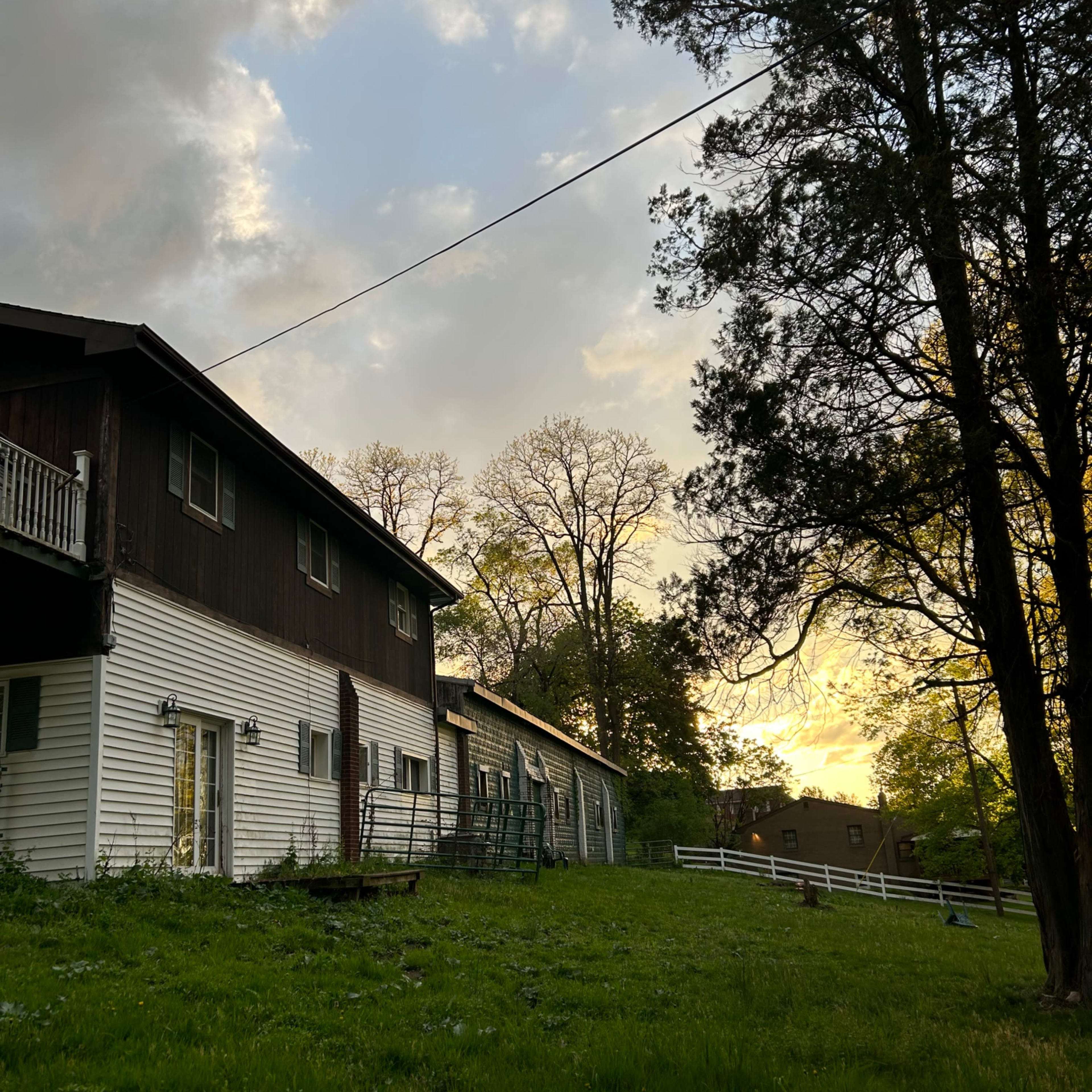 A large, two-story house with a dark wooden facade next to a grassy area and trees, under a cloudy sky during sunset.