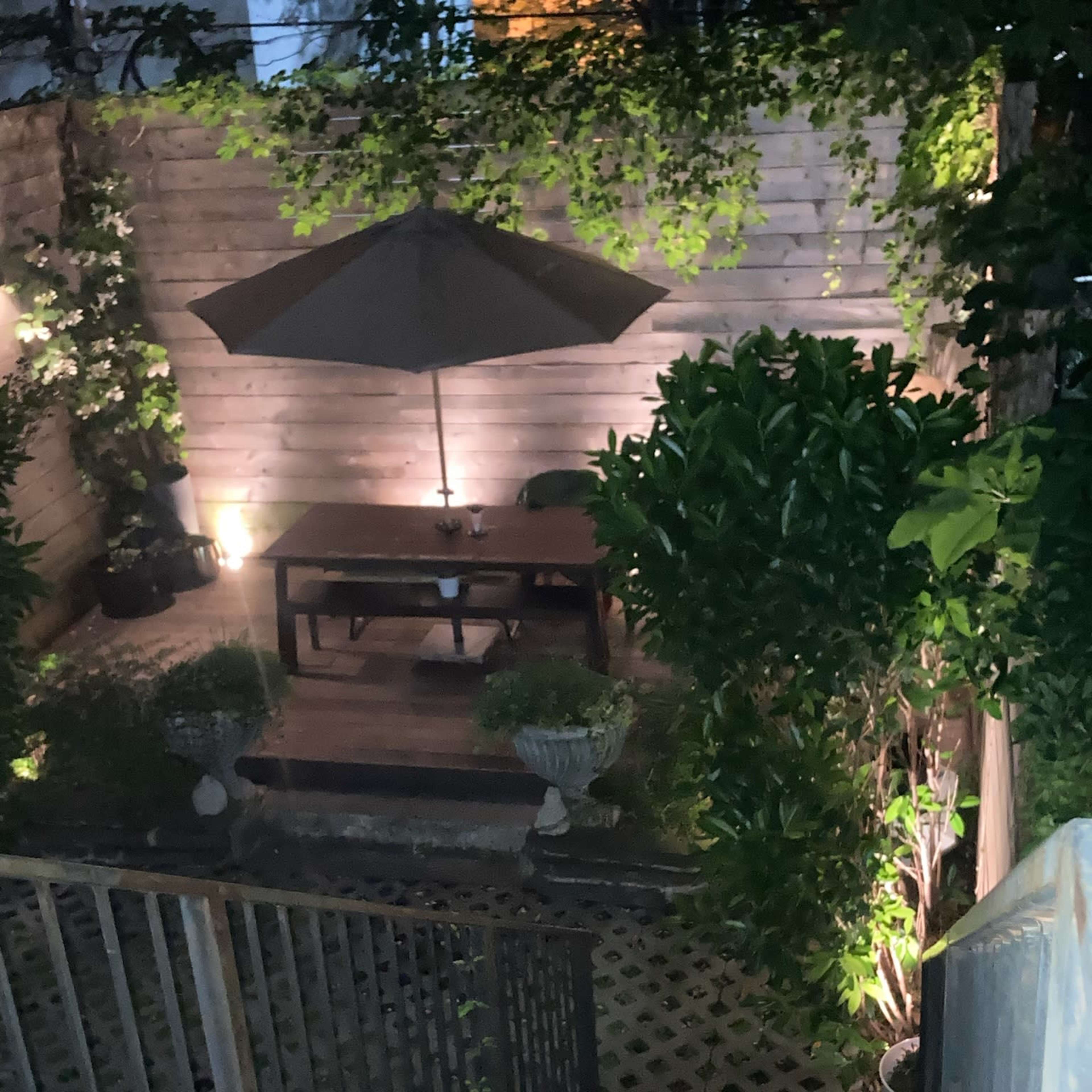 A wooden staircase leads down to a dimly lit backyard with a table and umbrella surrounded by plants.