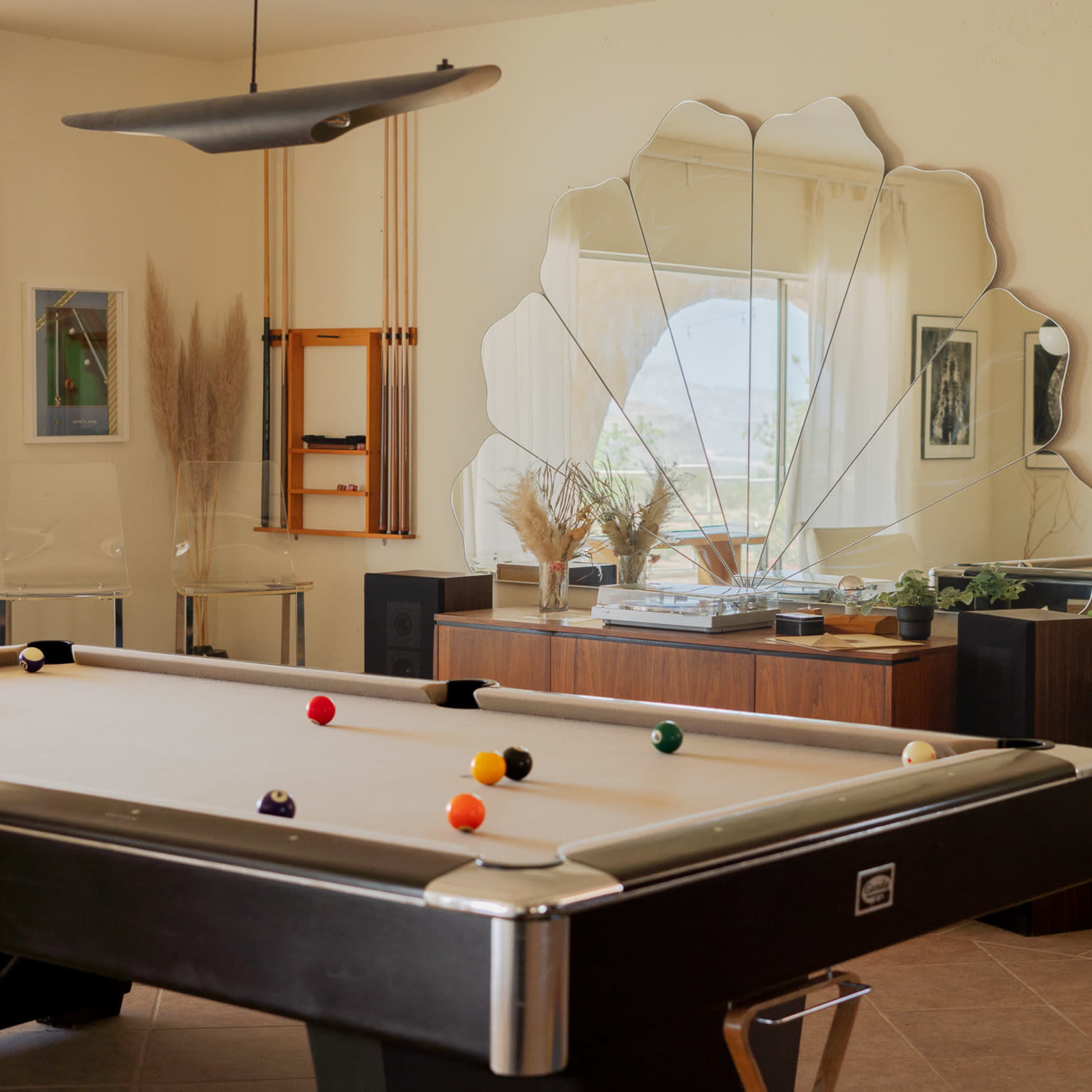 A pool table with balls scattered across it is positioned in a sunlit room featuring a large, decorative mirror and modern furnishings.