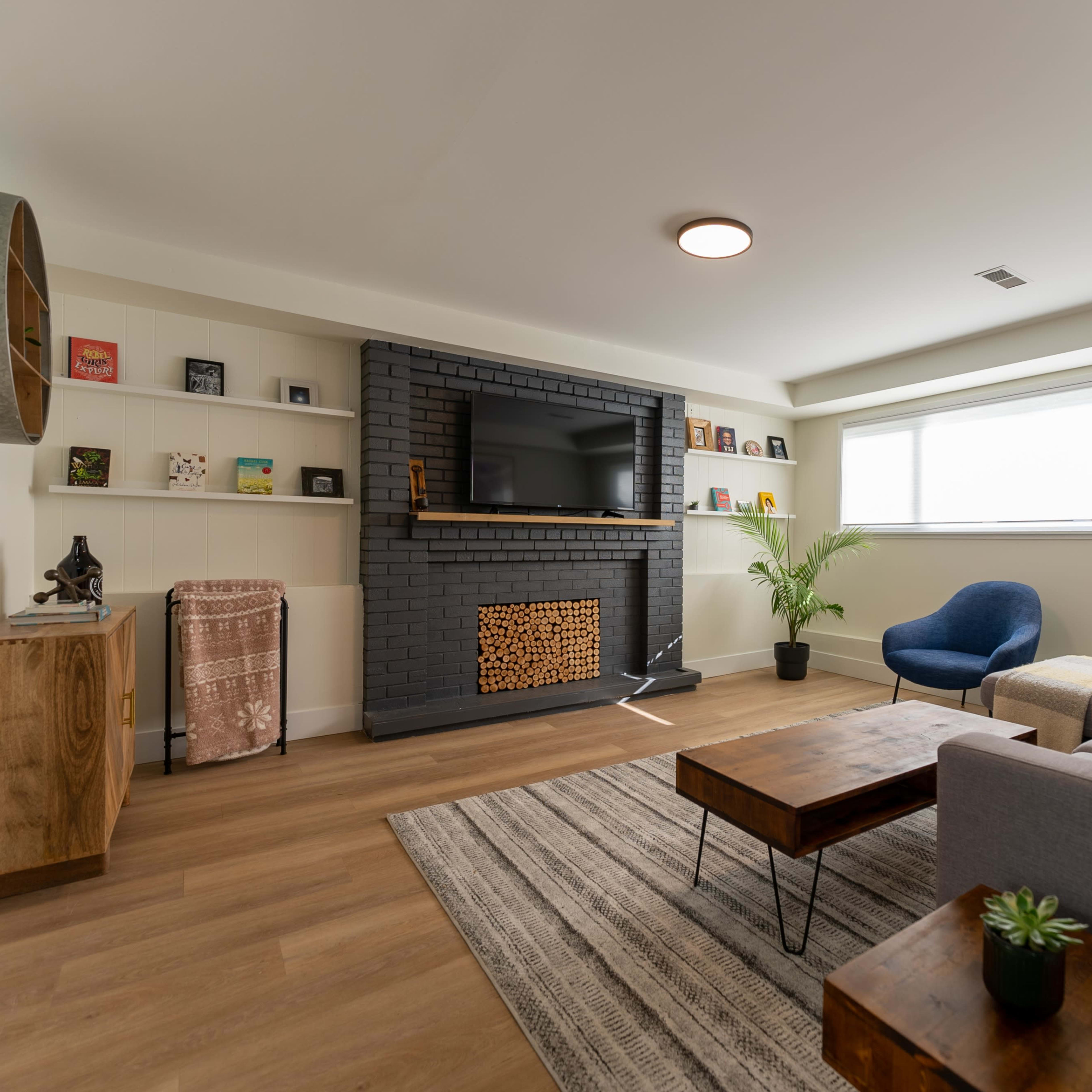 A modern living room with a gray sectional sofa, a wooden coffee table, a flat-screen TV mounted on a black brick wall, and a large window that provides natural light.