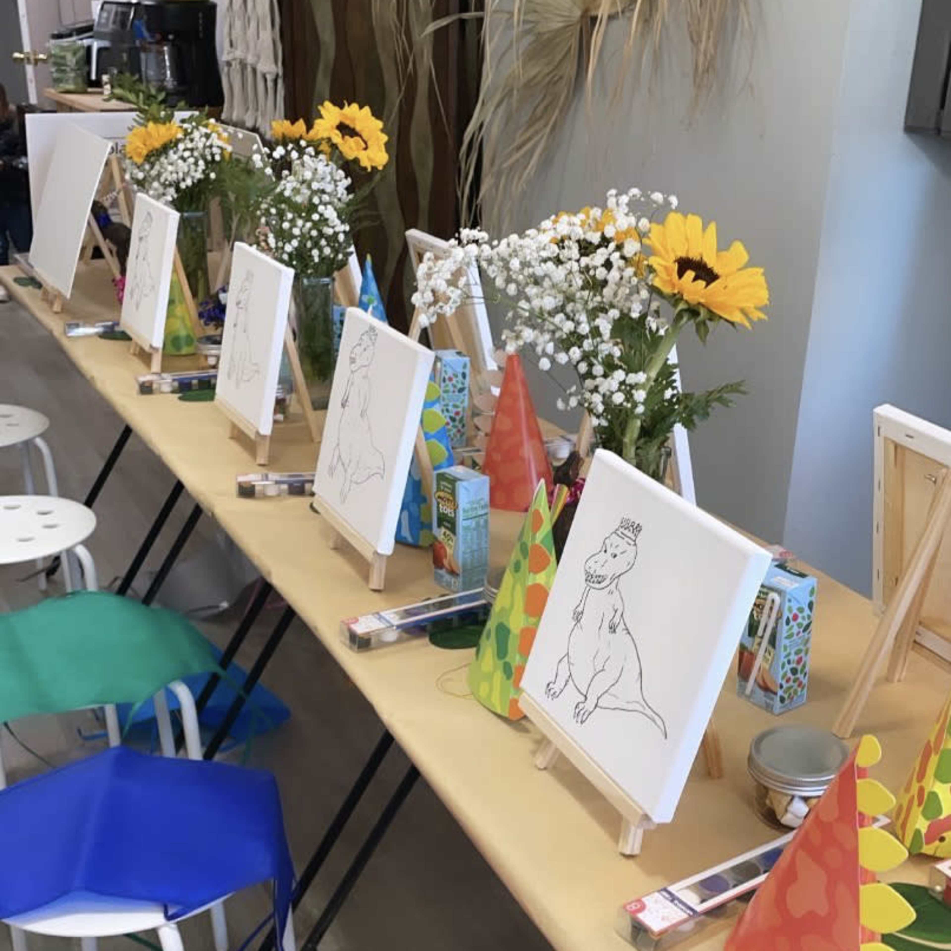 A long table is set up with easels holding blank canvases and colorful party hats surrounded by flowers and art supplies.
