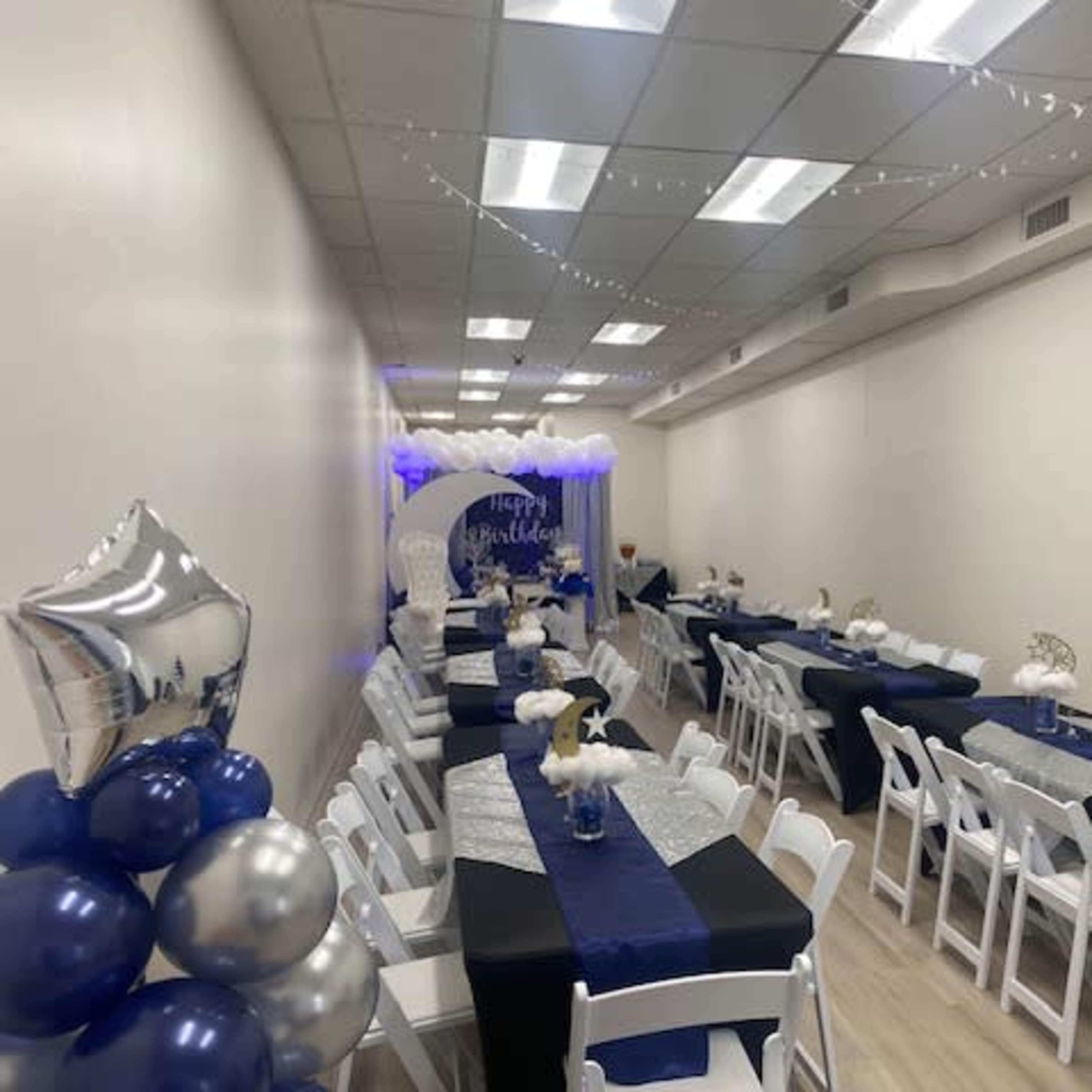 A decorated event space with several tables covered in black and silver linens, arranged to accommodate guests, and adorned with balloons and centerpieces.