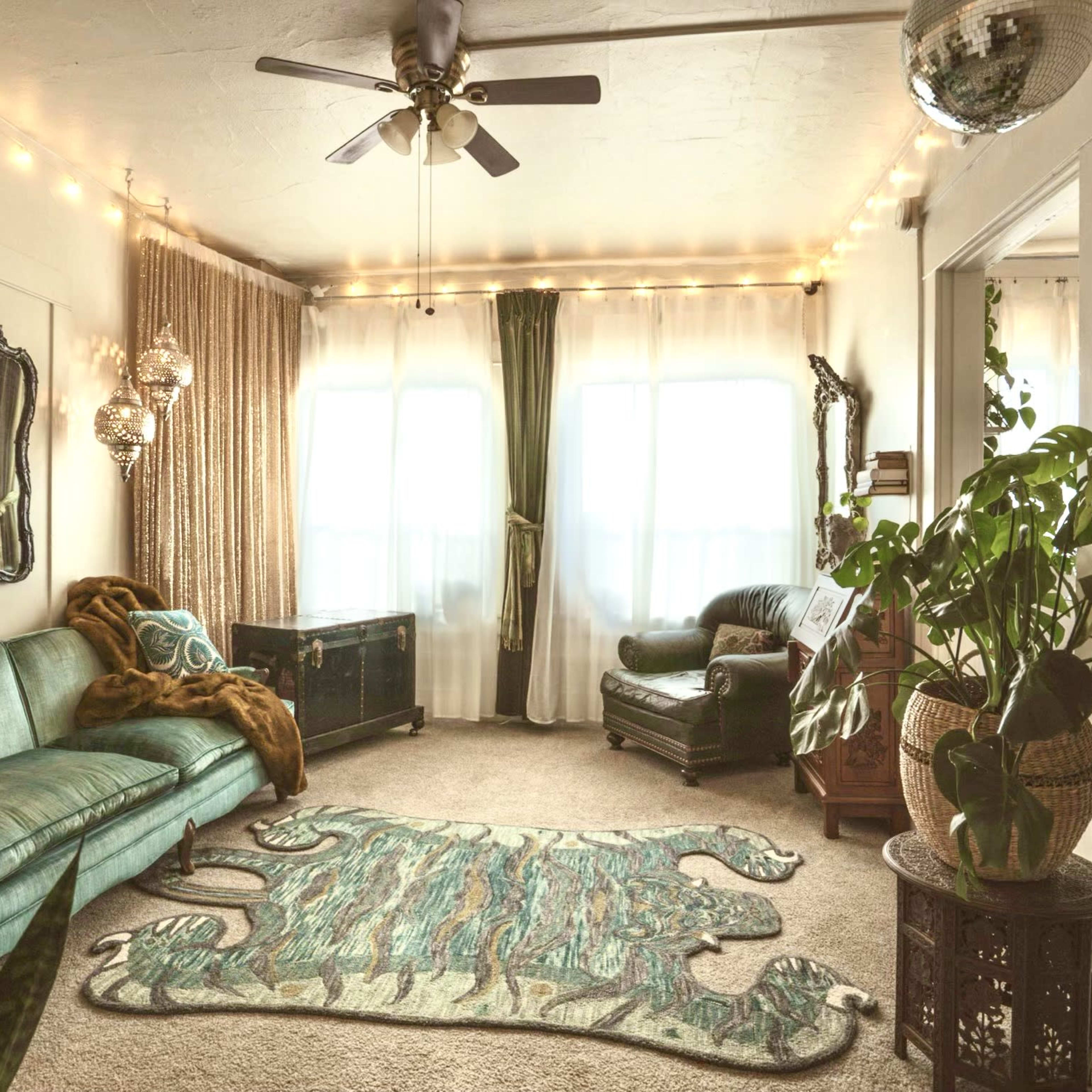 The scene features a cozy living room with a green sofa, patterned area rug, large windows, and decorative plants, illuminated by string lights.