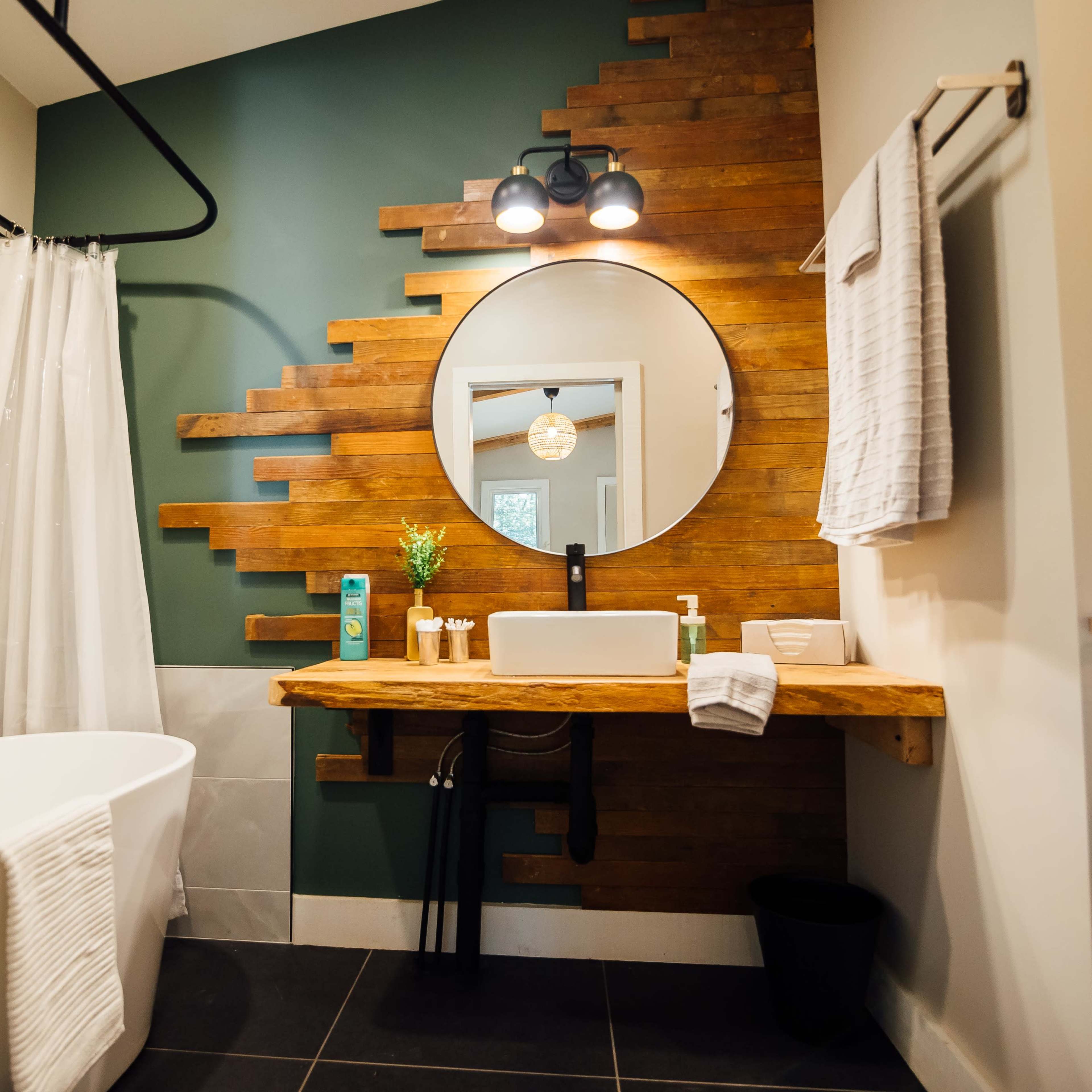 The bathroom features a modern design with a freestanding bathtub, a circular mirror, and a wooden sink countertop against a textured green wall.
