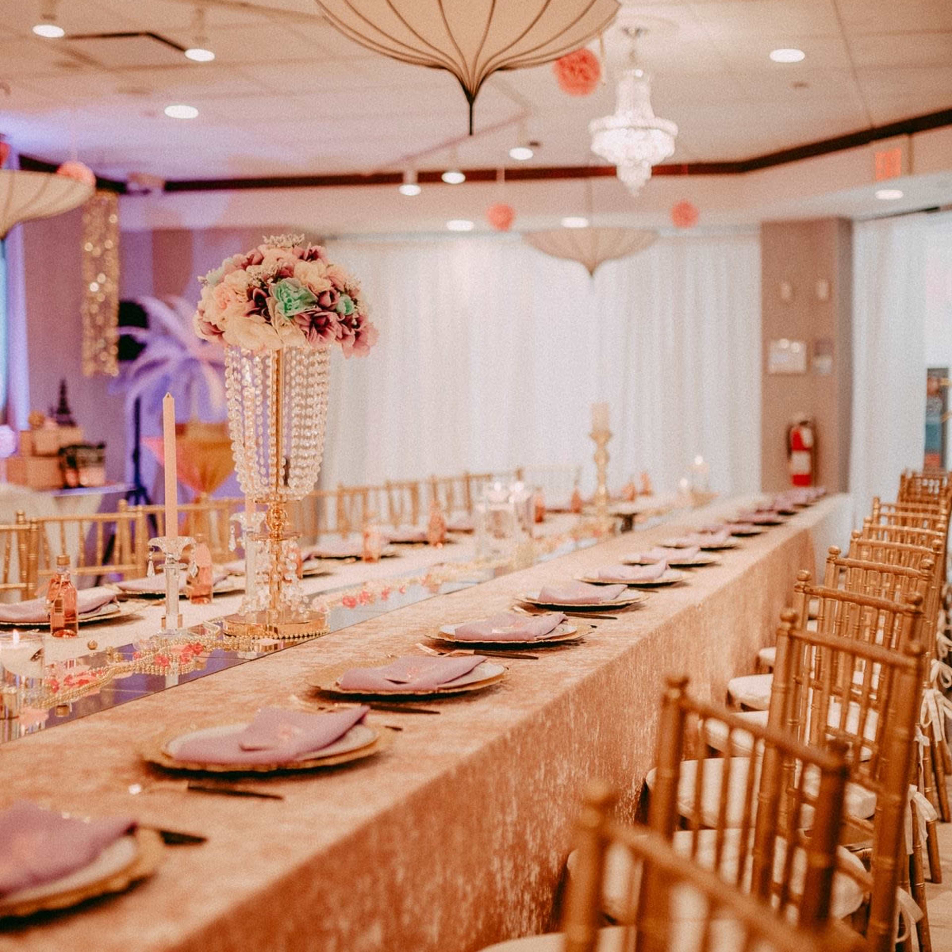 A long banquet table is set with elegant place settings, adorned with a decorative centerpiece and draped in a sparkling cloth under stylish lighting fixtures.