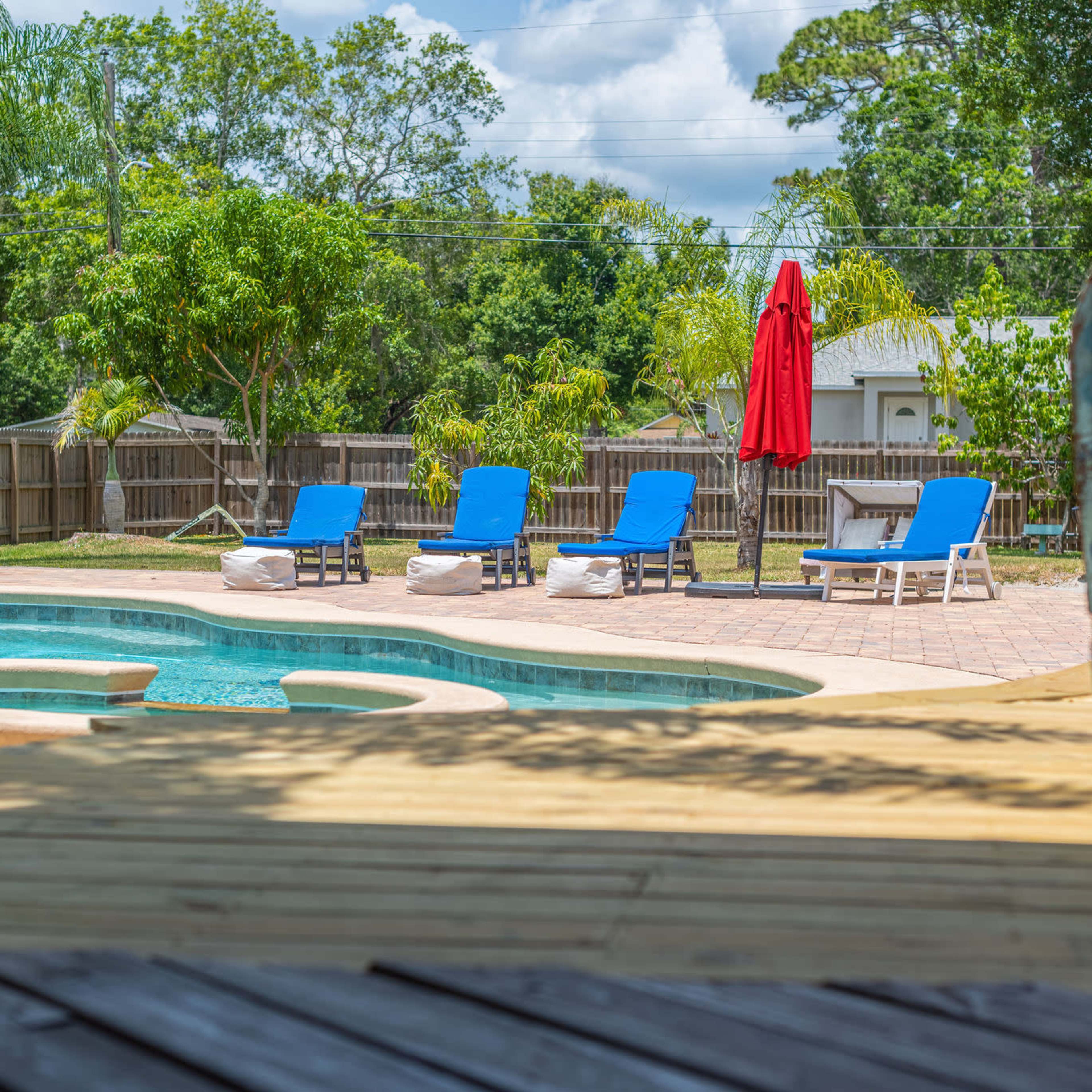 A swimming pool with lounge chairs and a red umbrella is surrounded by greenery in a fenced backyard.