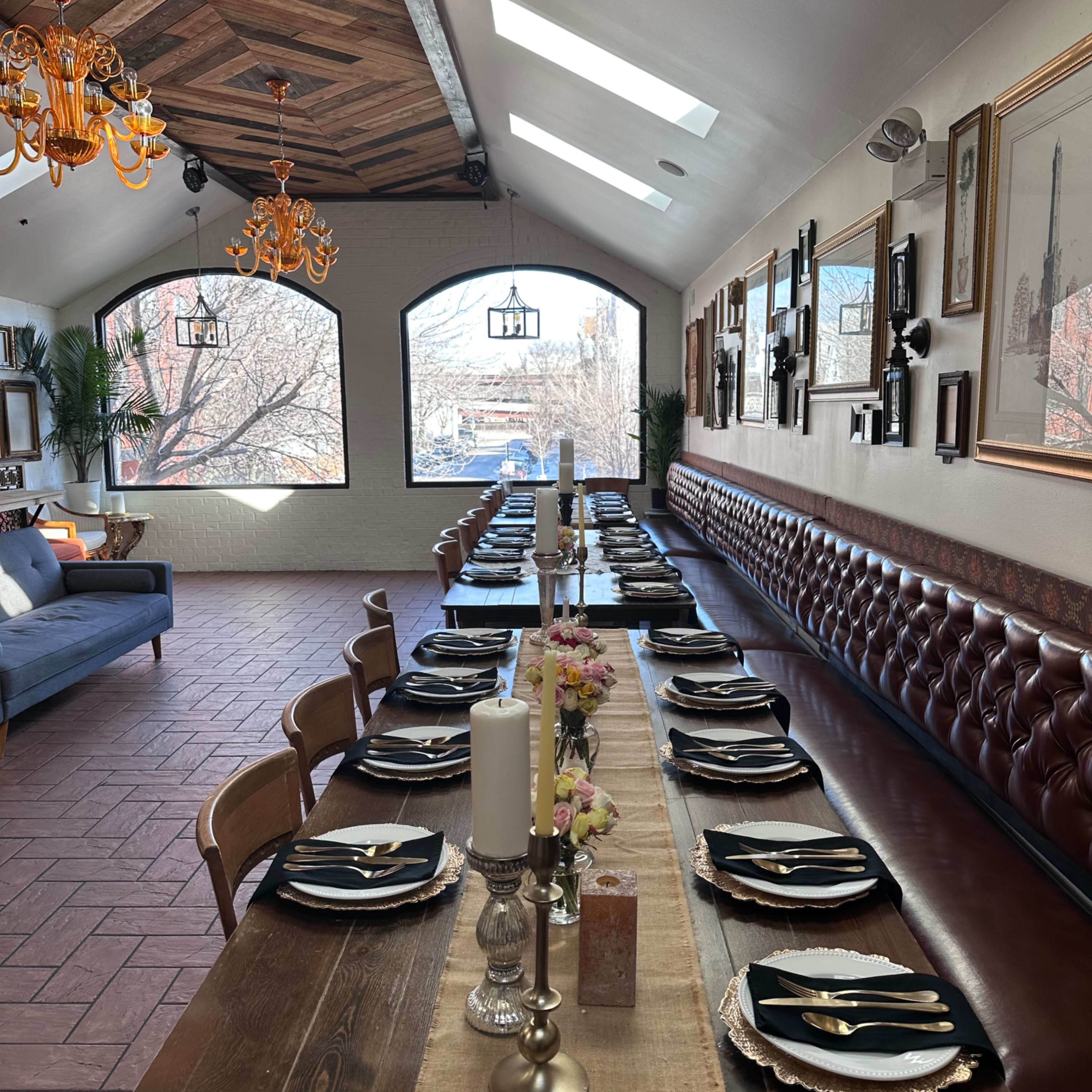 A long, elegantly set dining table with black plates and silverware is arranged in a well-lit room featuring large windows and a wooden ceiling.