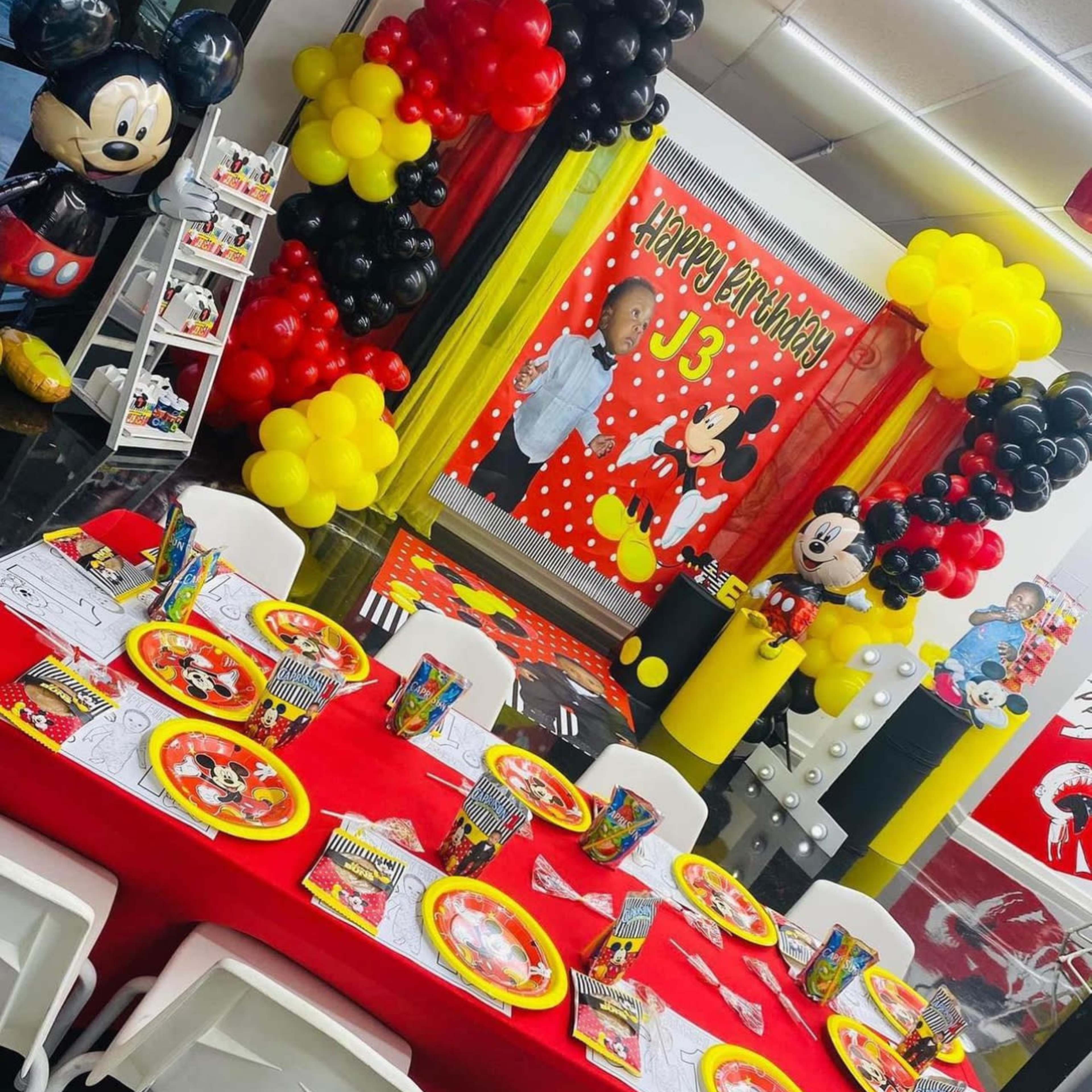 A festive birthday party setup features a table decorated with red tablecloths, yellow and black balloons, and Mickey Mouse-themed plates and decorations.