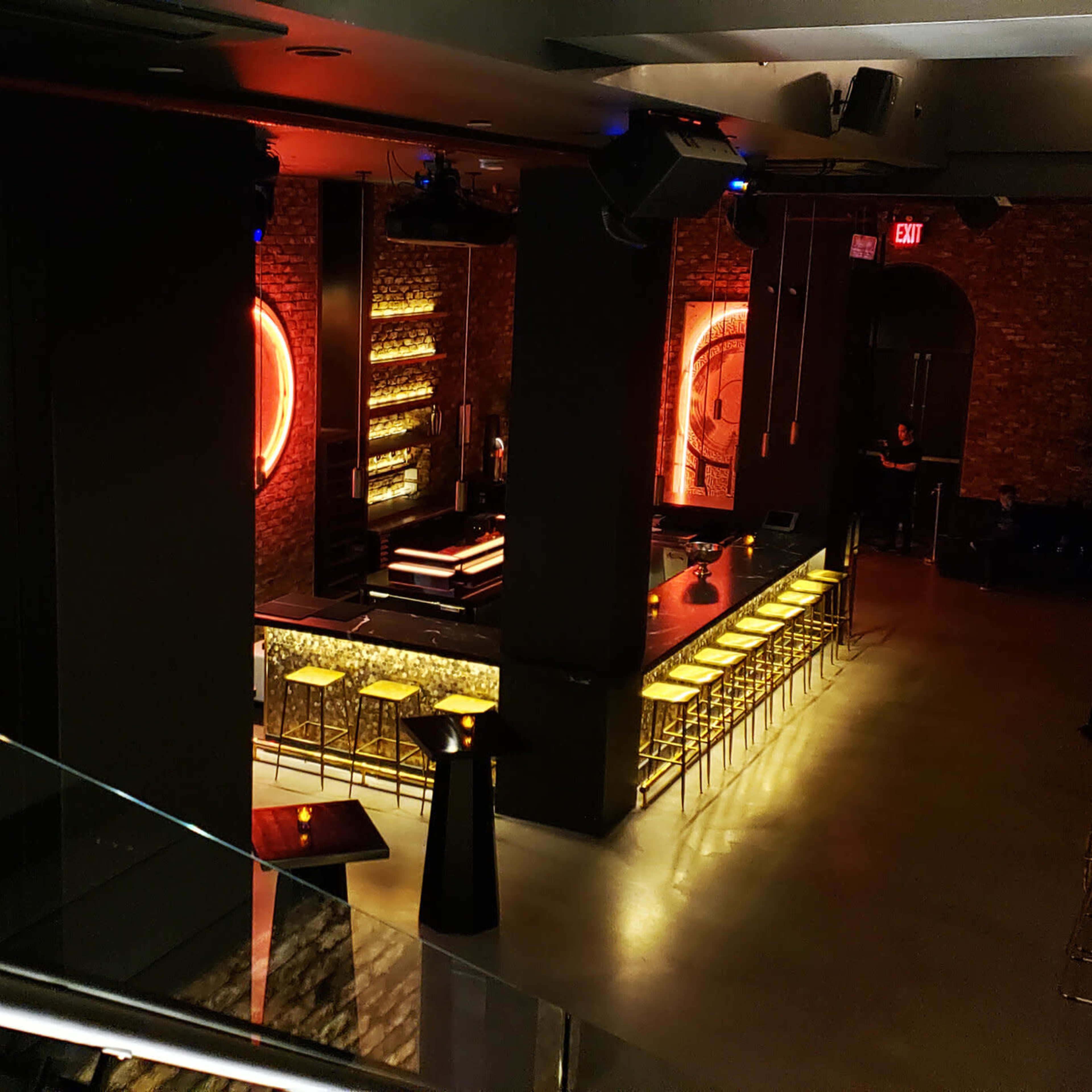 A dimly lit bar area with a neon-lit backdrop, featuring a long row of gold bar stools and a display screen on the wall.