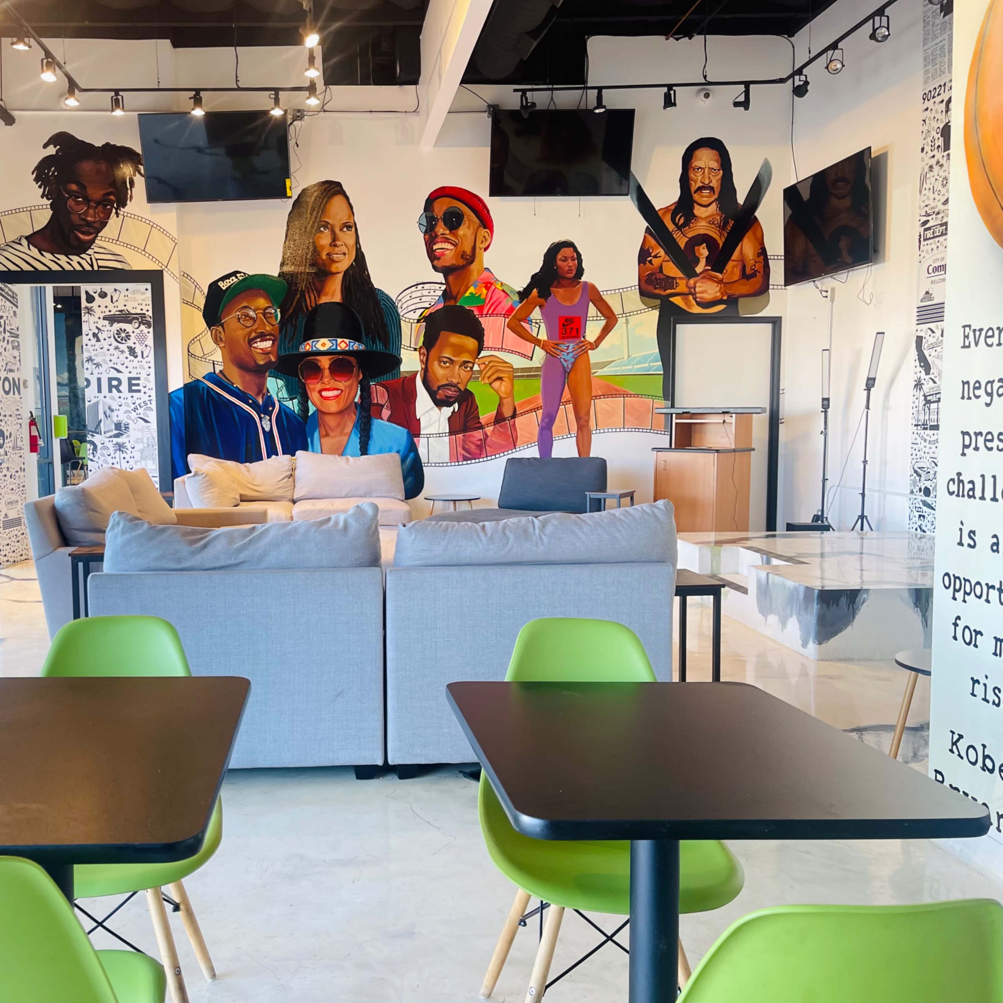 A modern cafe interior with a large mural featuring various characters on the wall behind a sofa and tables with green chairs.