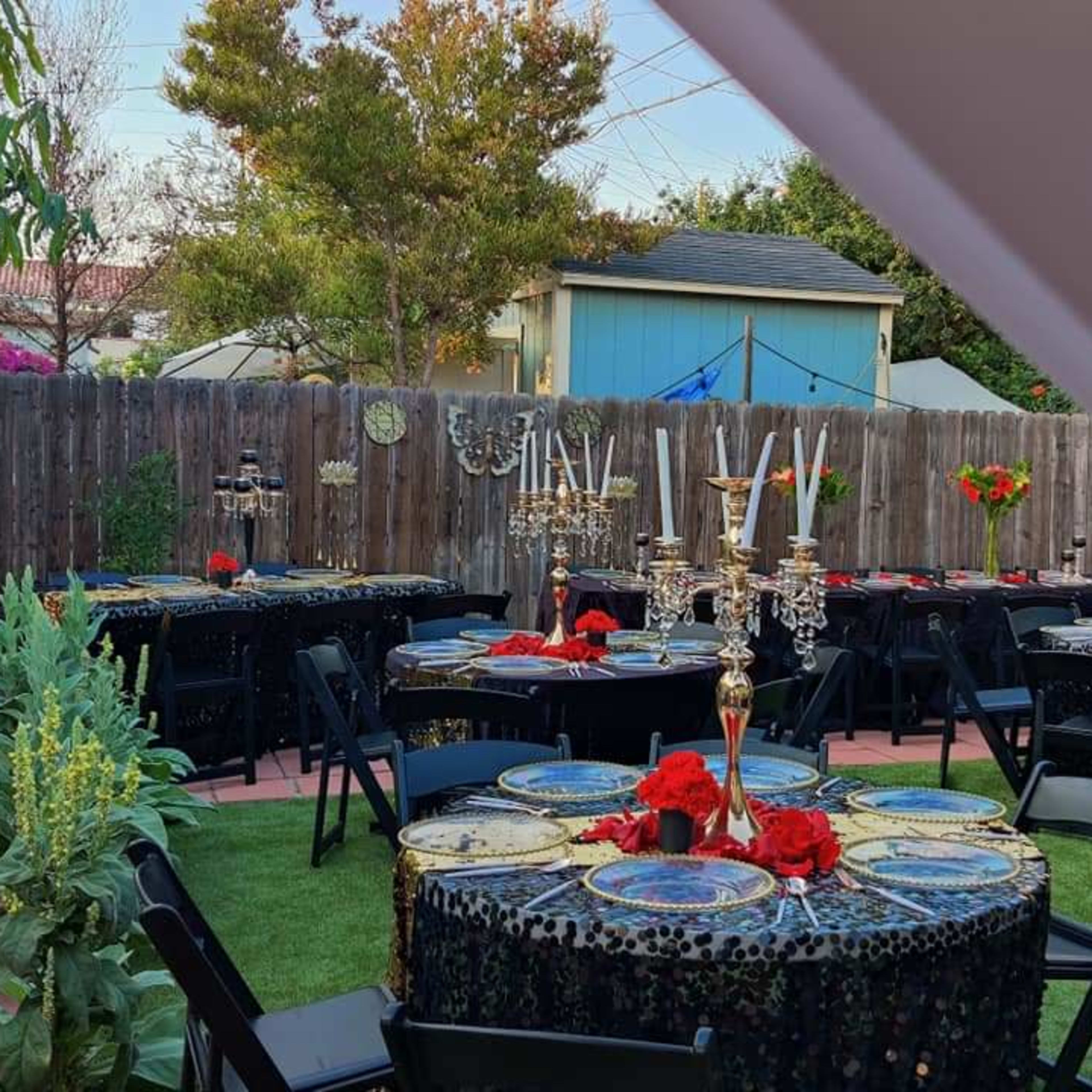 A backyard is set up for a party with several elegantly decorated tables featuring black tablecloths, floral centerpieces, and candelabras.