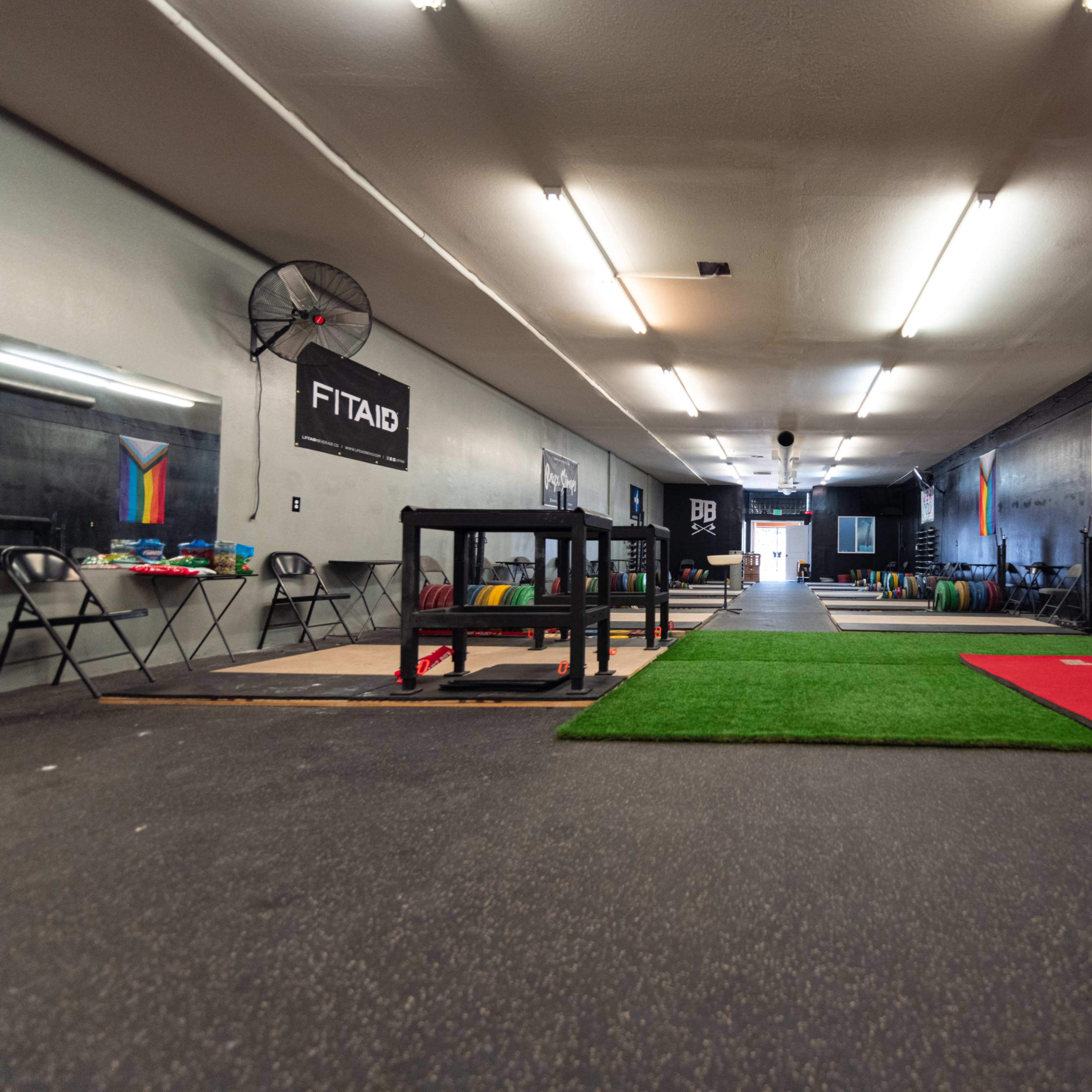 The interior of a fitness gym with a long hallway, exercise equipment on the sides, and various flooring surfaces including artificial grass and mats.