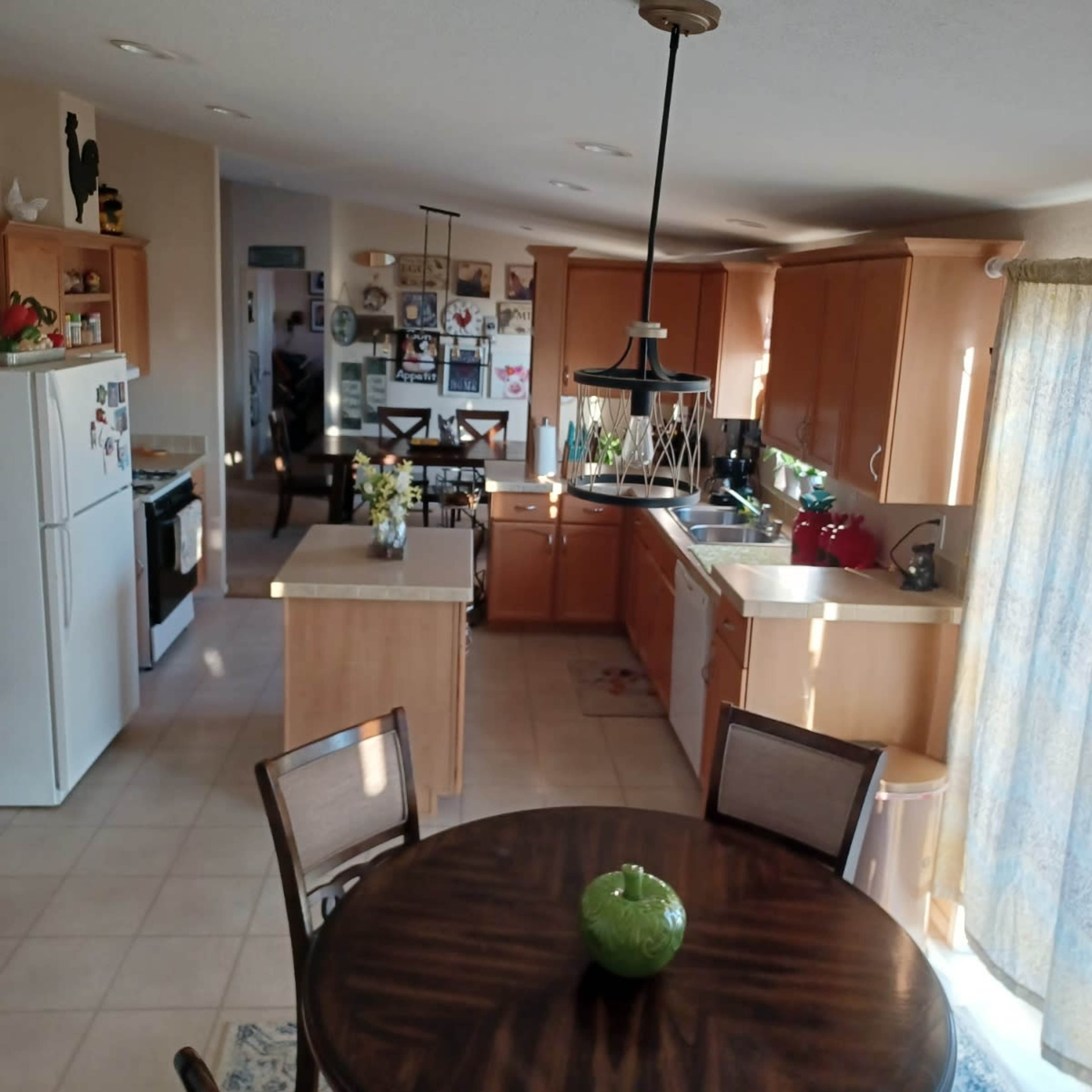 A kitchen with wooden cabinets, an island, and a dining table set for four, featuring a curtain and various decorations on the walls.