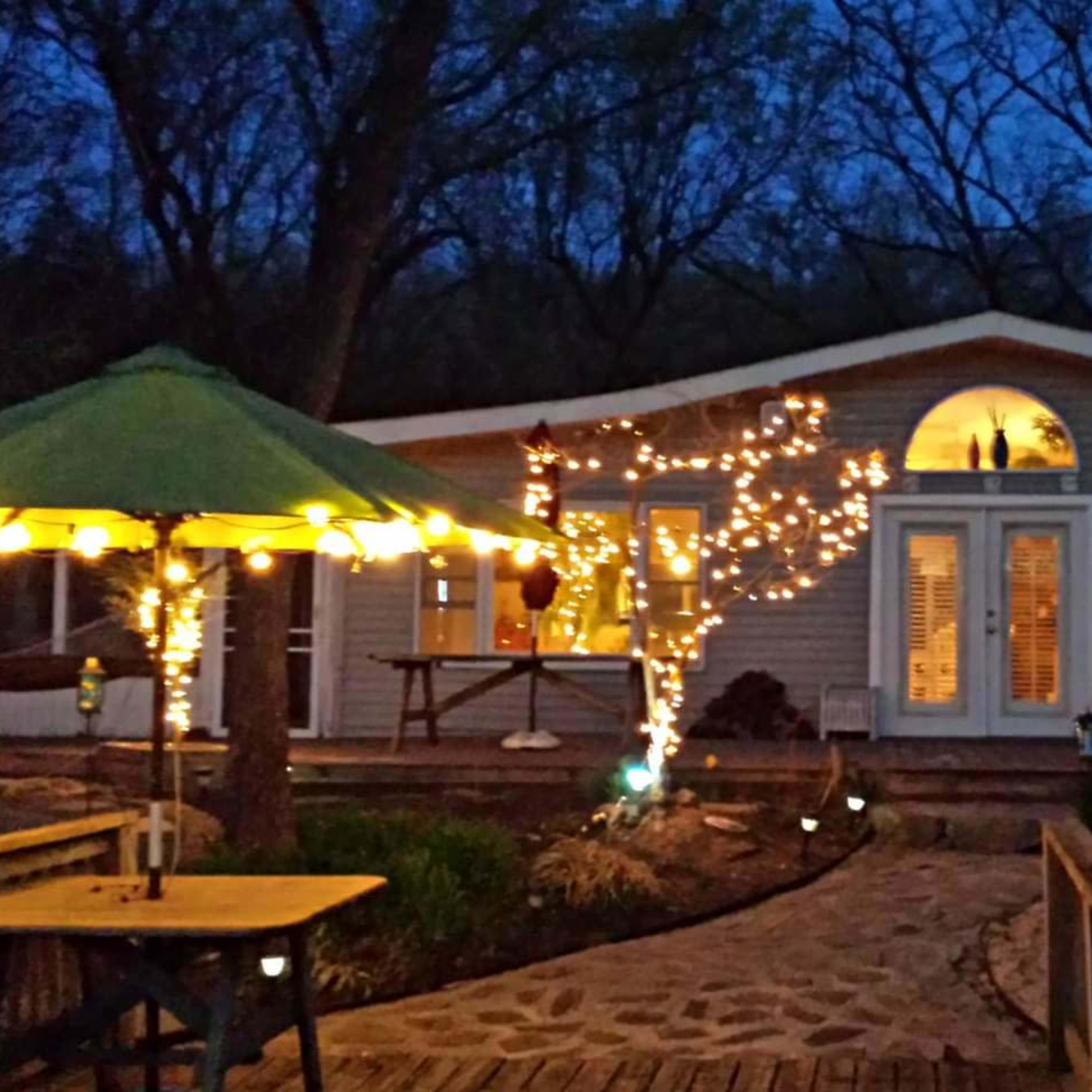 A cozy house with string lights wrapped around a tree is nestled in a wooded area at dusk, with a green umbrella-covered table in the foreground.