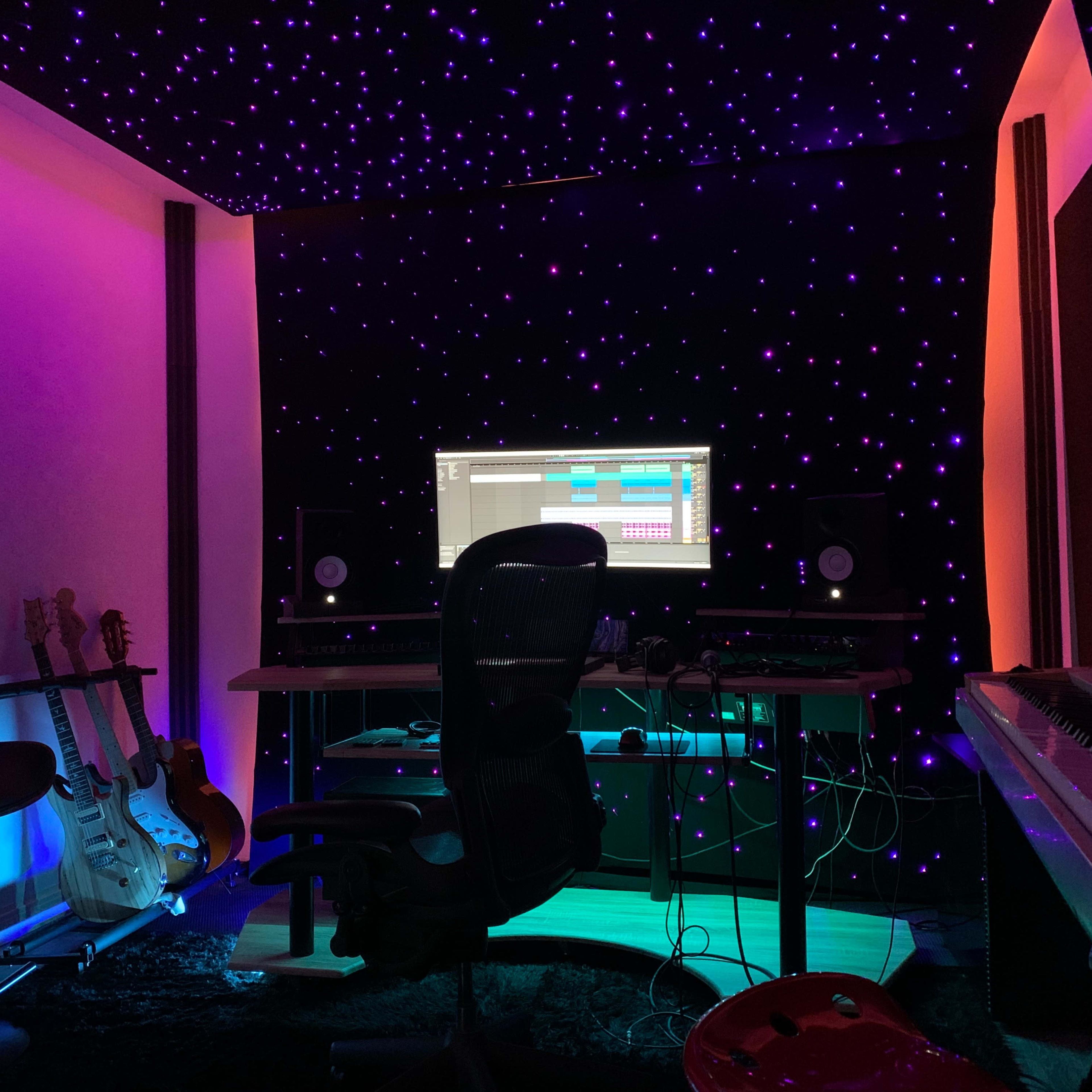 A music studio features a computer setup with monitors, surrounded by guitars and illuminated by colorful lights against a starry backdrop.