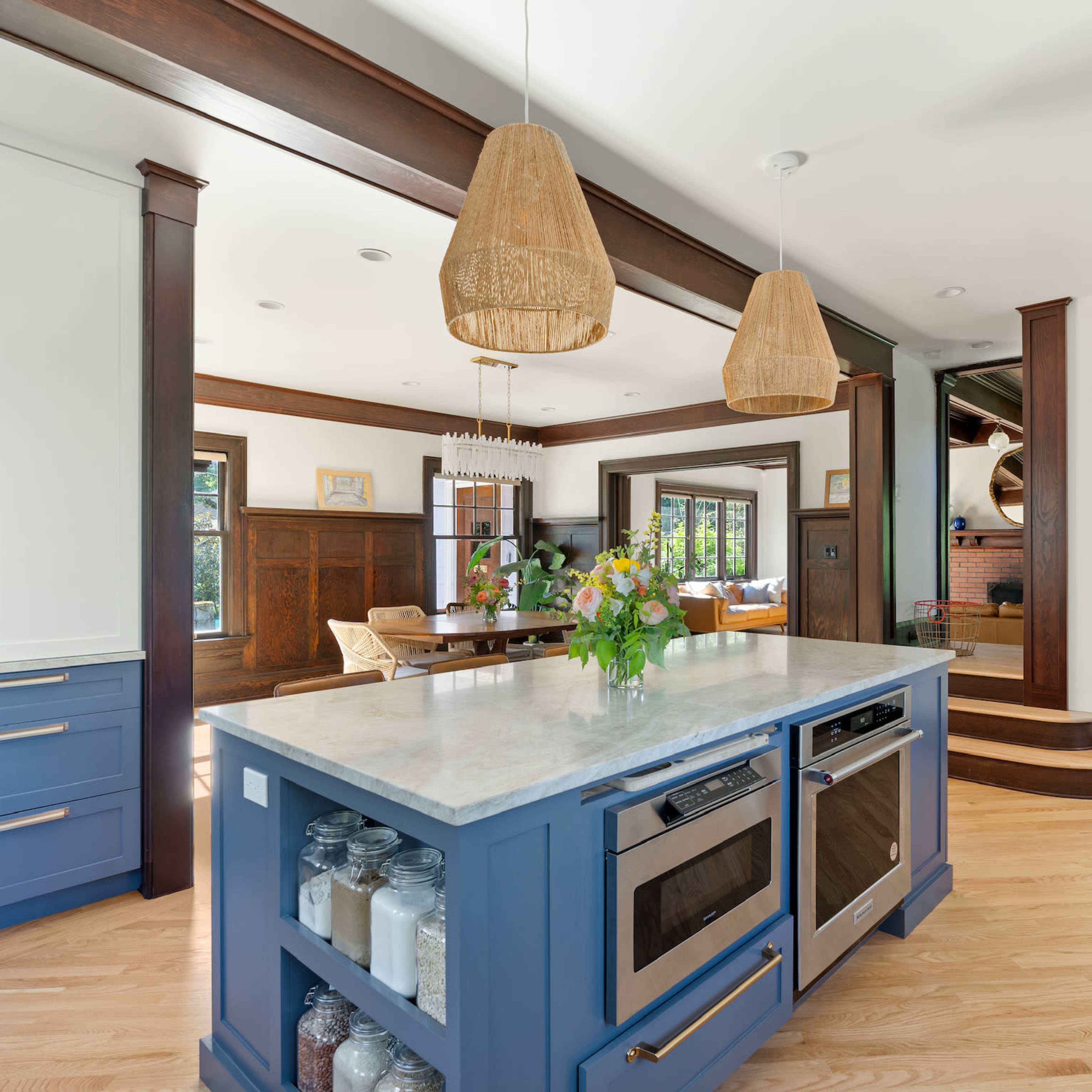 A modern kitchen features a blue island with a marble countertop, open shelves displaying jars, and pendant lights hanging above.