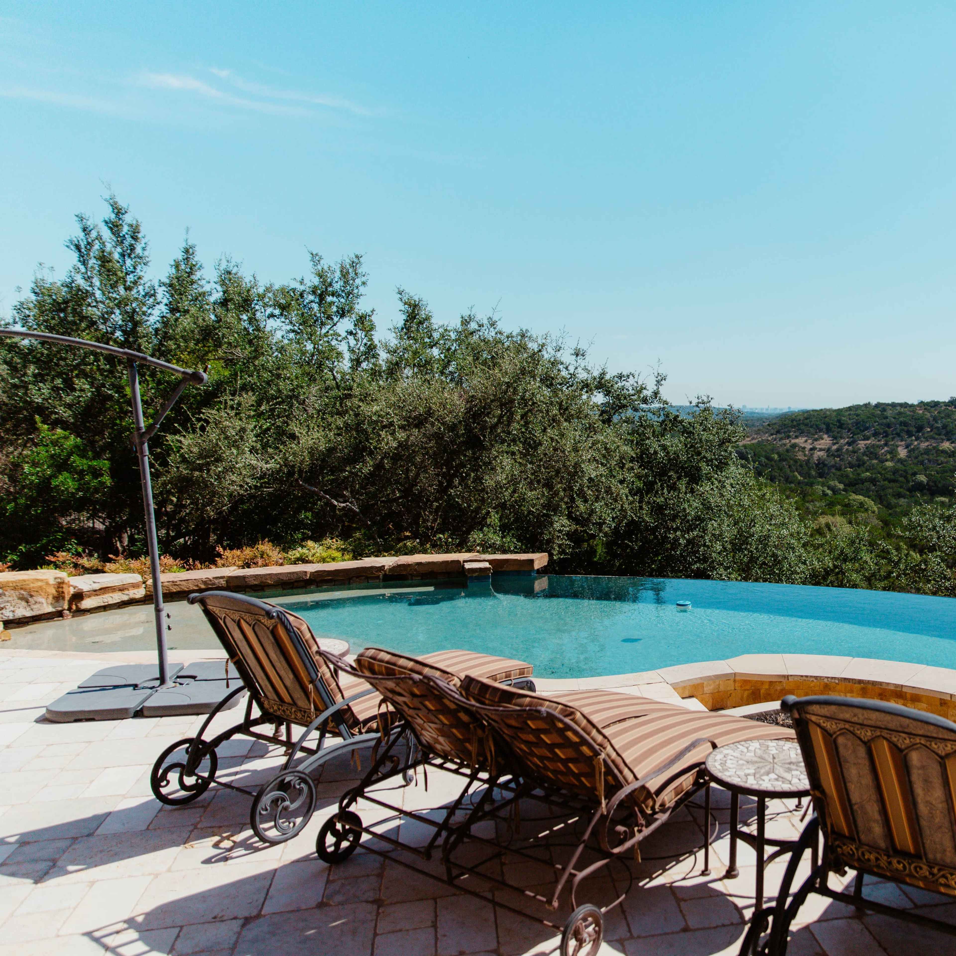 The scene features an infinity pool with three lounge chairs positioned beside it, overlooking a lush, hilly landscape under a clear blue sky.