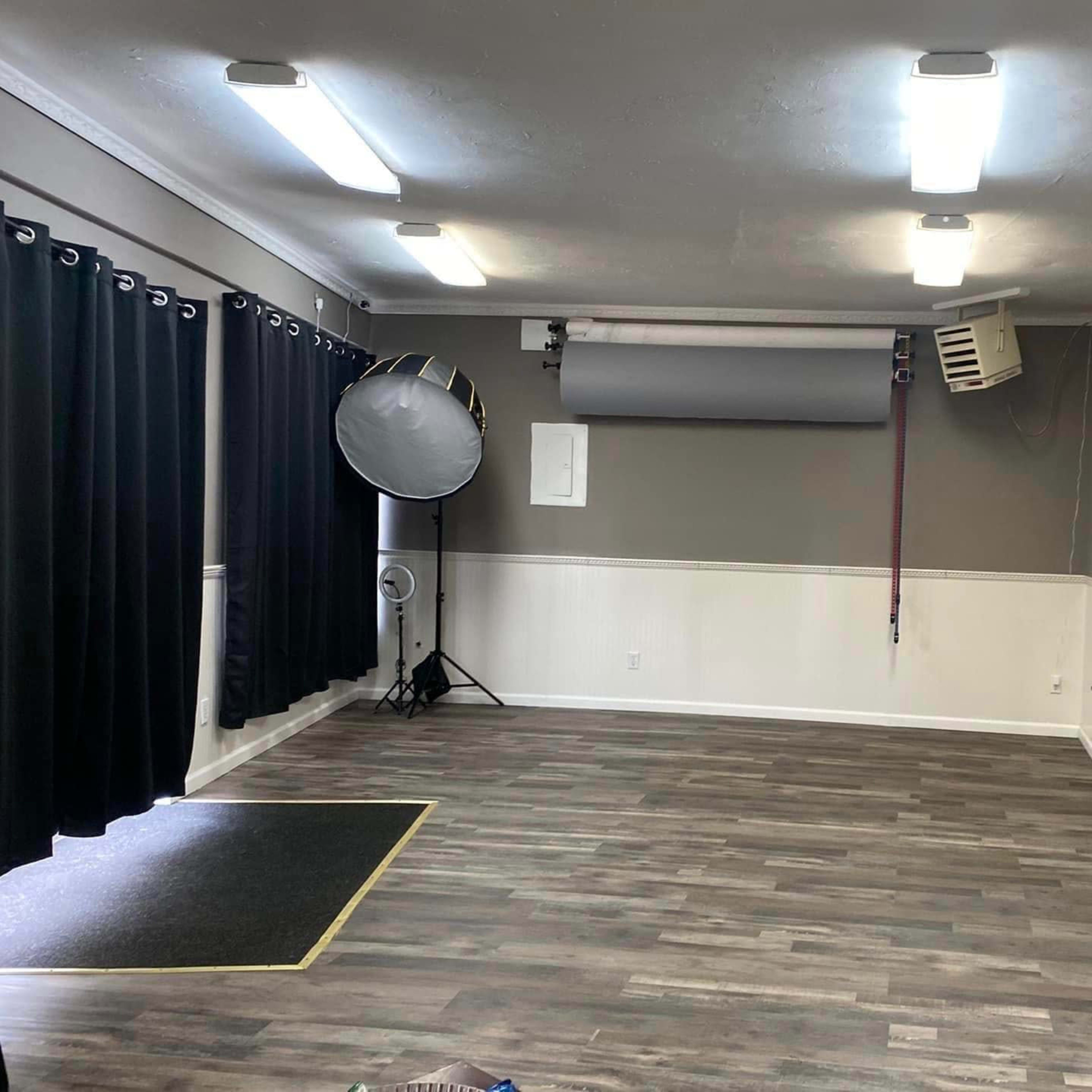 A spacious, empty studio with gray walls, wooden flooring, a large reflector, and black curtains on the windows.