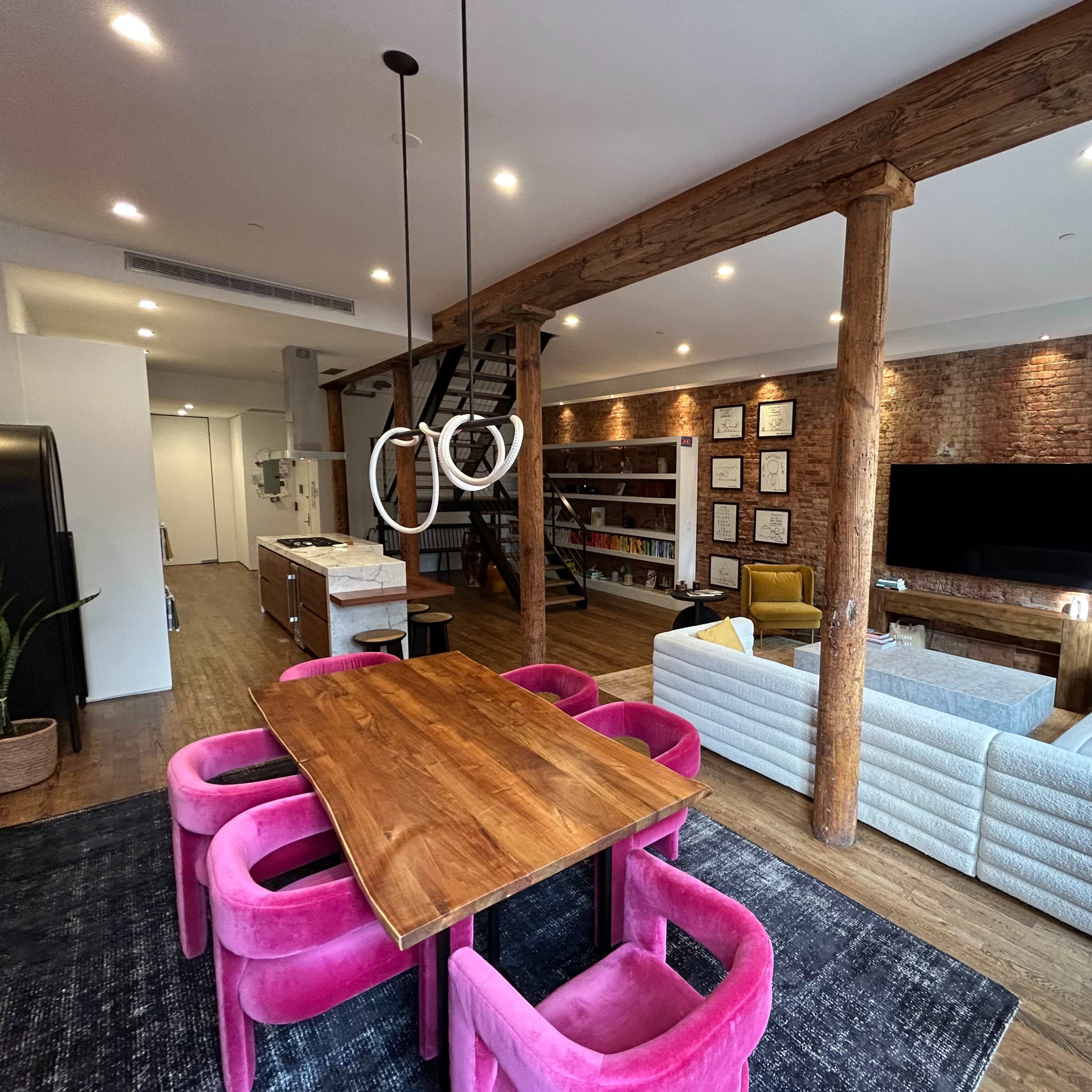 A modern open-concept living space featuring a dining area with a pink table and chairs, a cozy living area with sofas, and wooden beams supporting the ceiling.