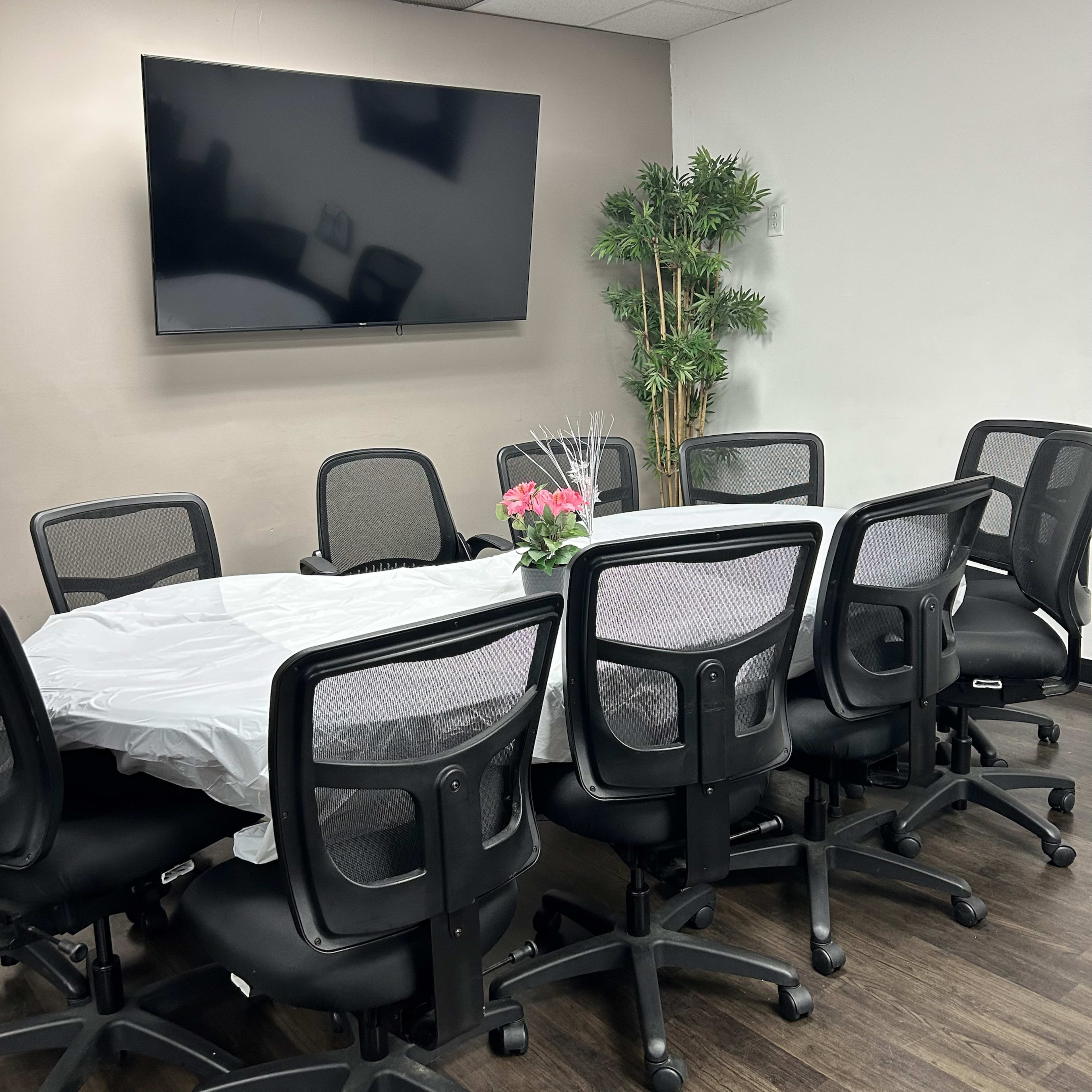 A conference room features a long table surrounded by office chairs, equipped with a large wall-mounted television and a potted plant.